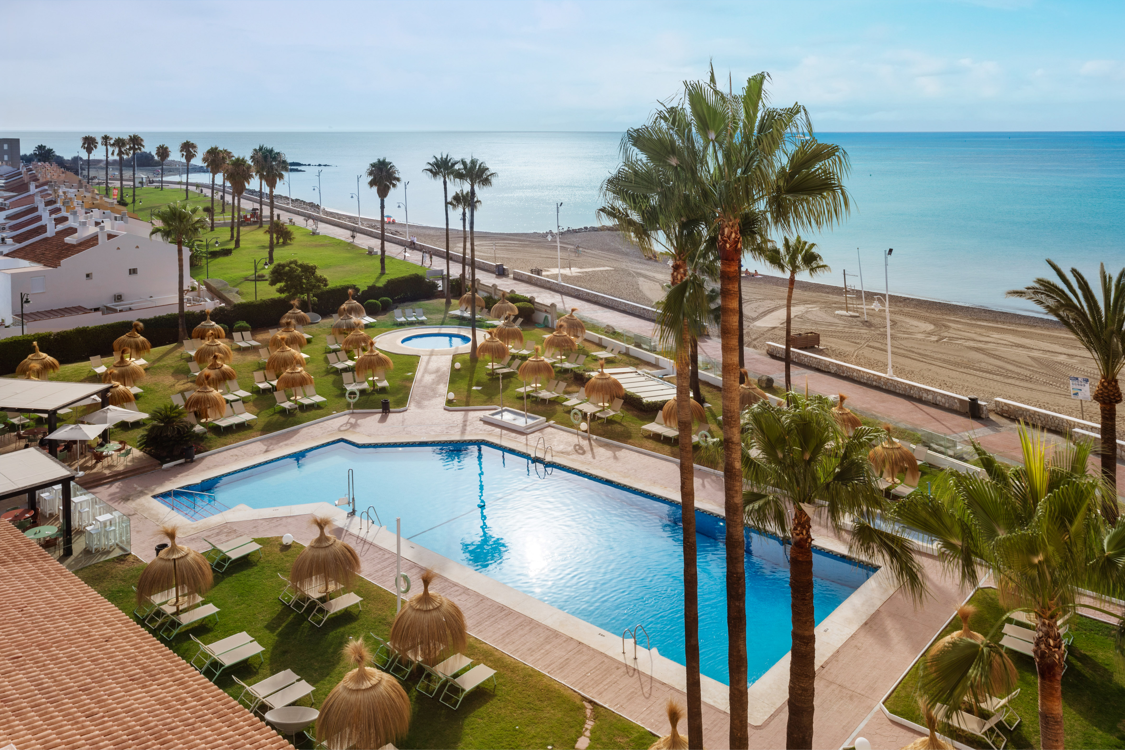 Coastal resort with large pool, palm trees, sun loungers, and sandy beach.