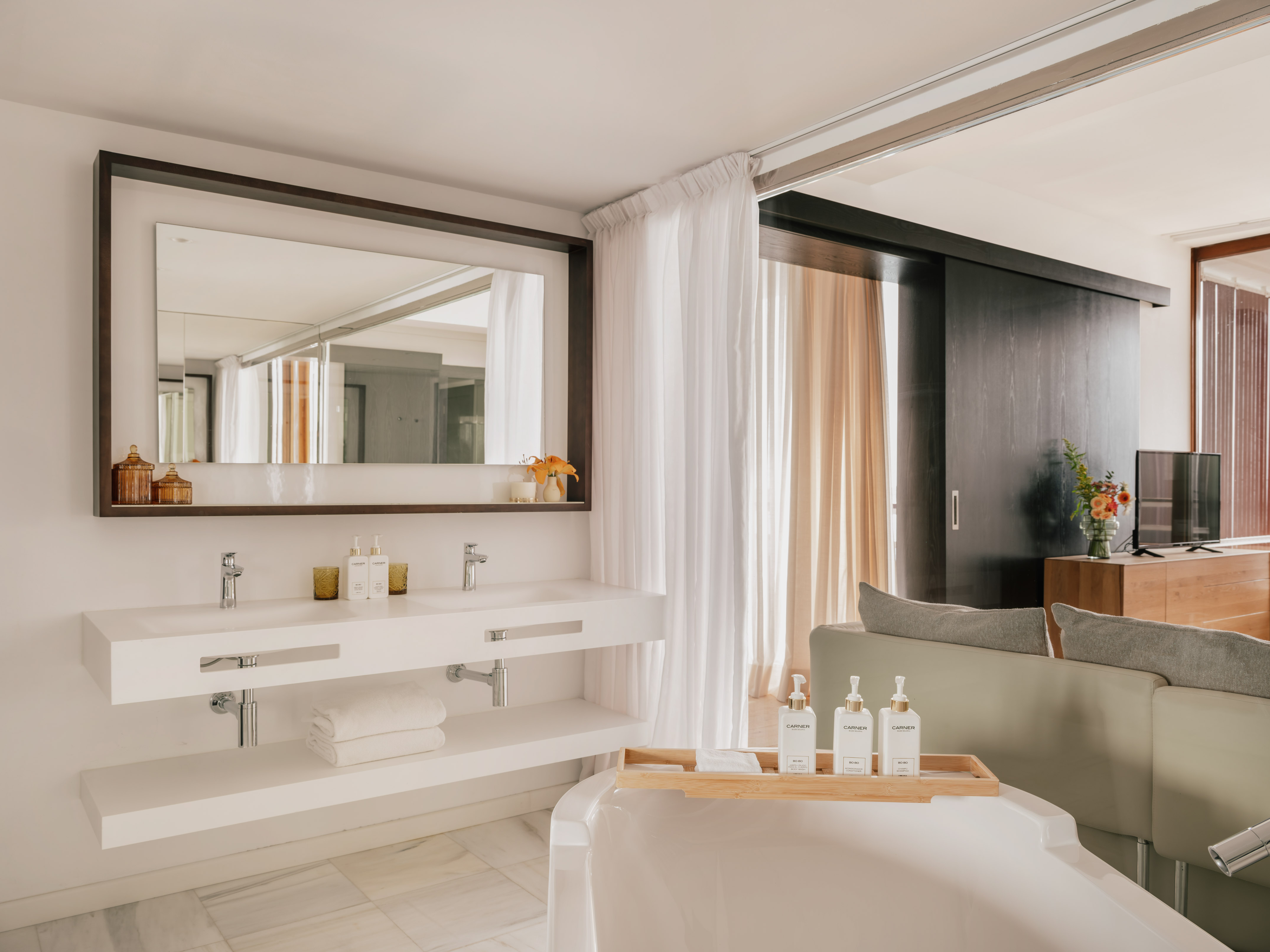 Modern hotel bathroom with double vanity, freestanding tub, and living area.