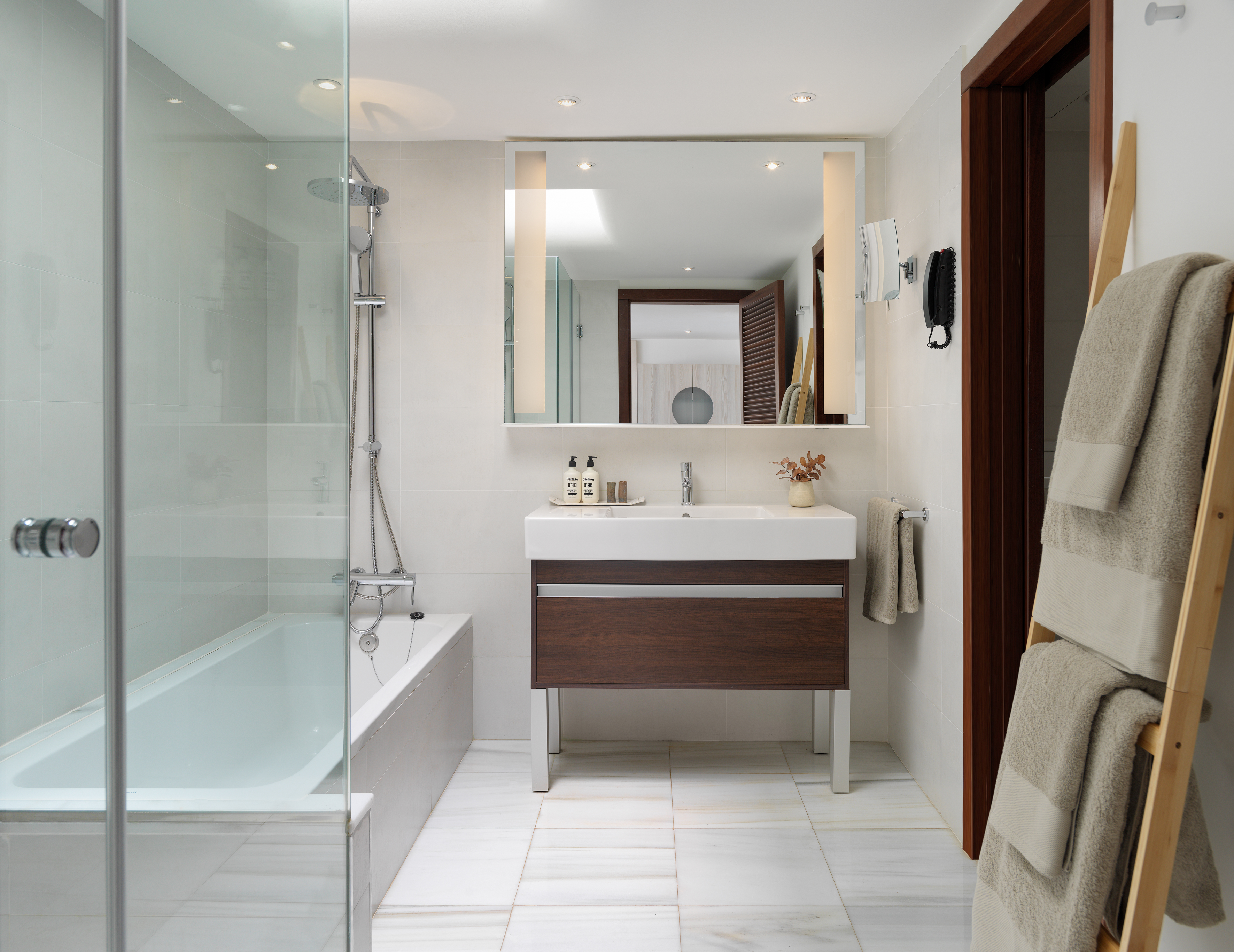 Modern hotel bathroom with shower-tub combo, wood vanity, and illuminated mirror.