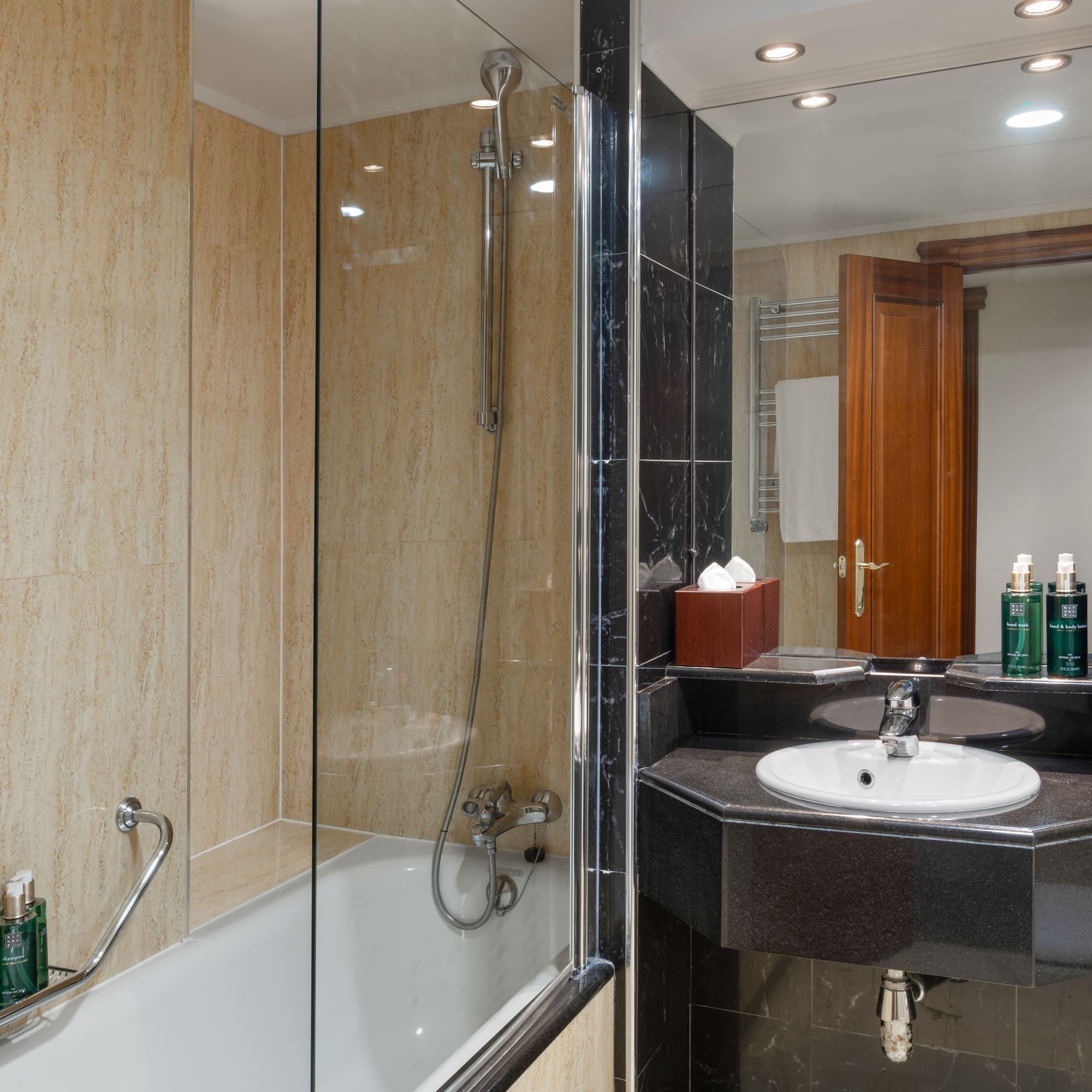 Modern hotel bathroom with shower, bathtub, black marble vanity, beige tiles.