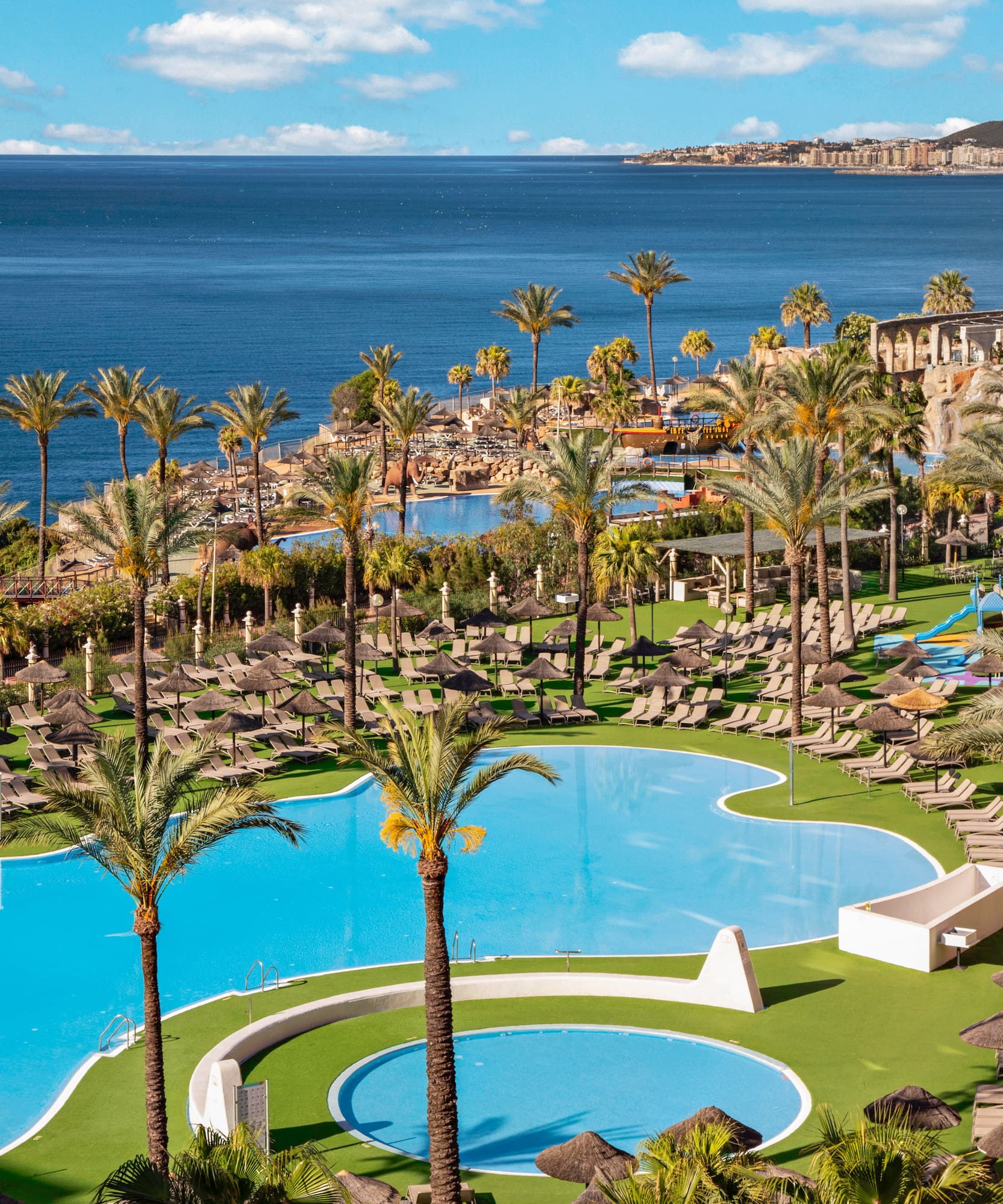 Tropical resort with pools, palm trees, ocean view, and sun loungers.