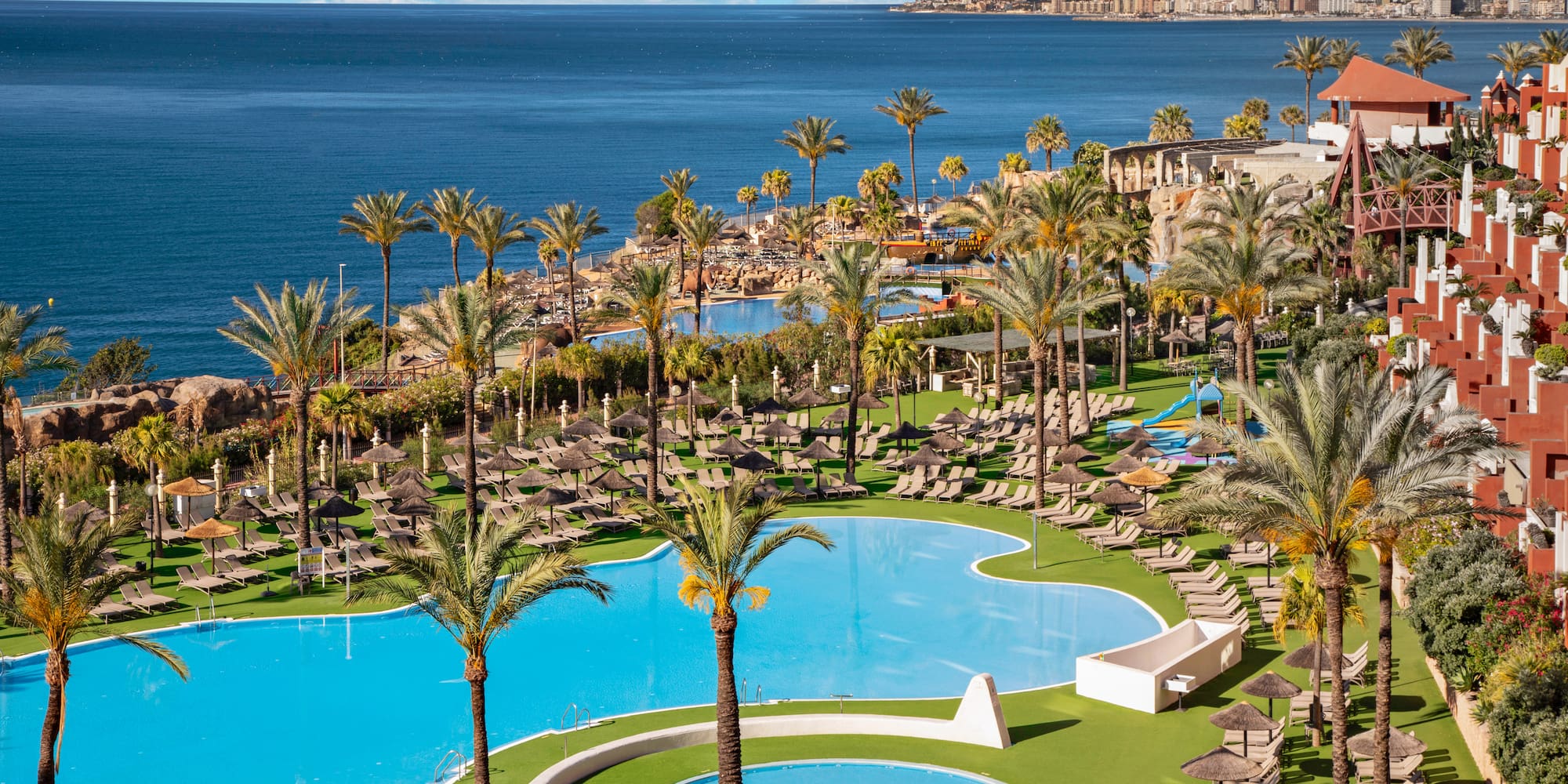 Hotel resort with large swimming pools, palm trees, ocean, and coastal city.