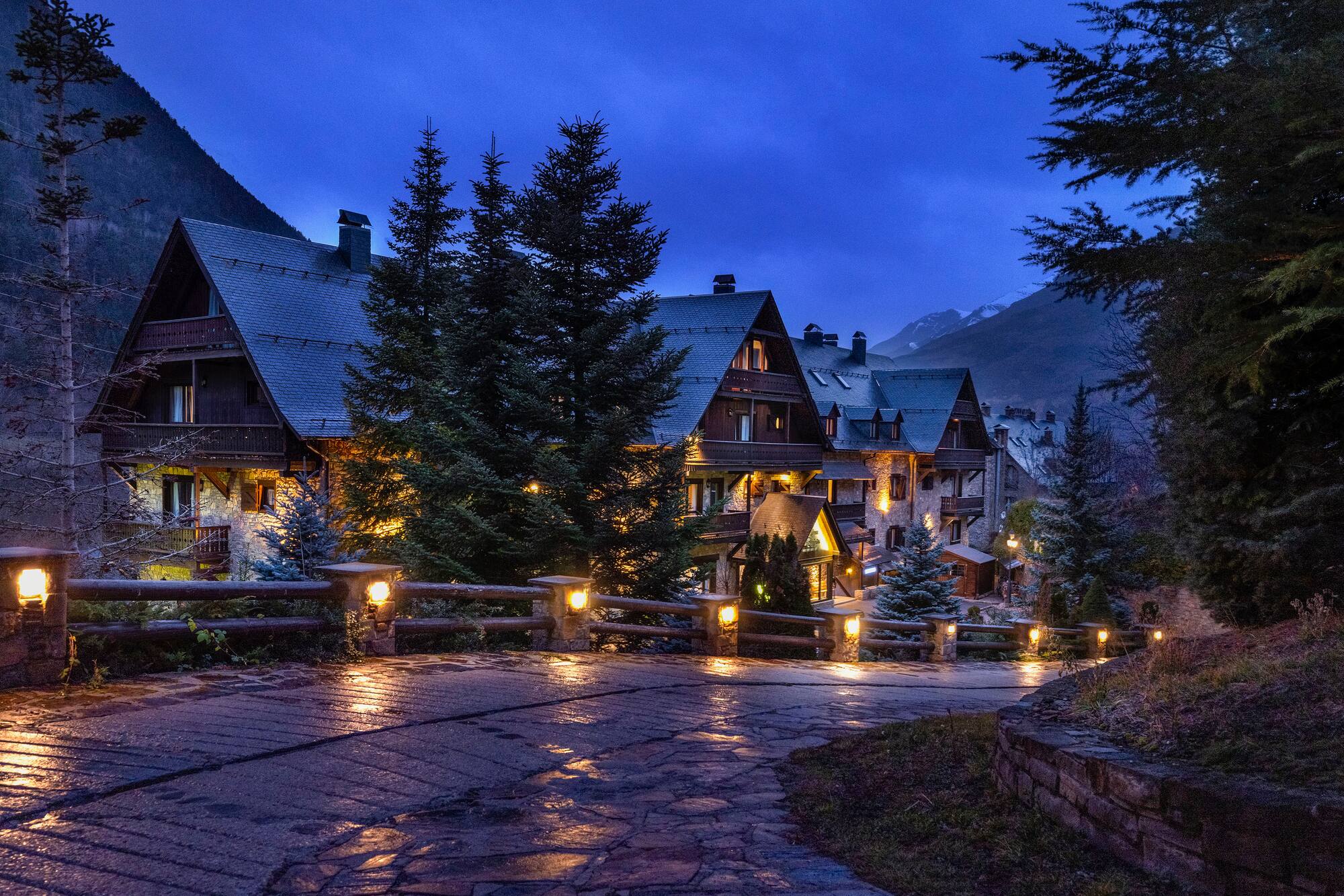 Mountain hotel lit up at dusk, surrounded by pine trees and distant peaks.