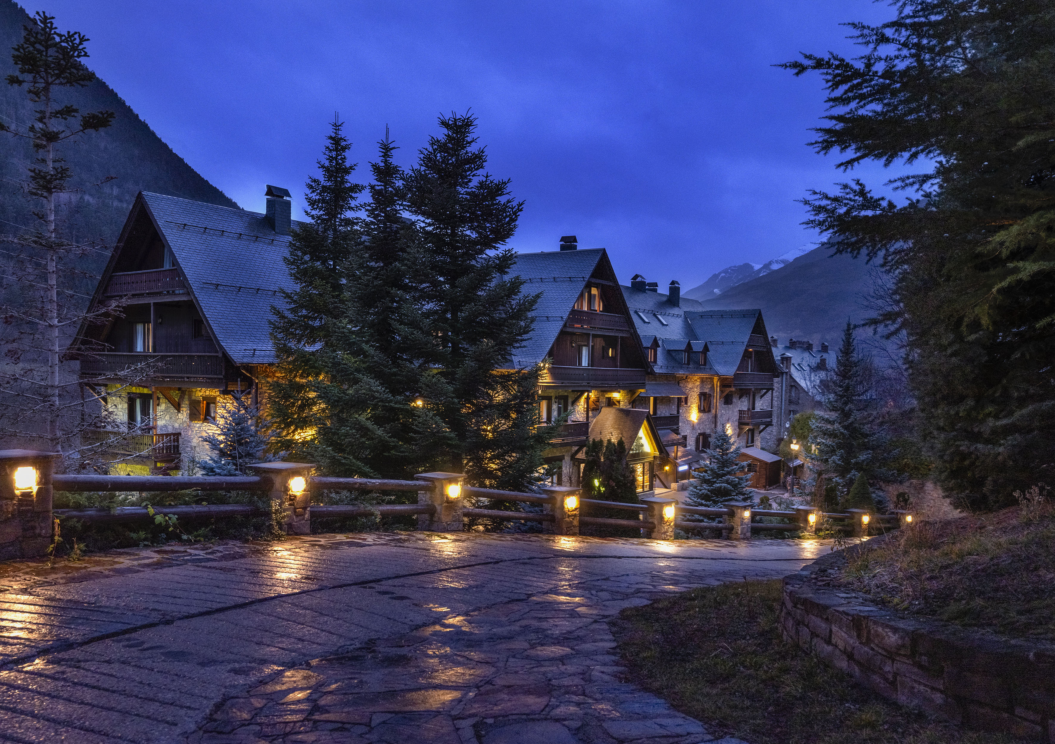 Mountain hotel lit up at dusk, surrounded by pine trees and distant peaks.