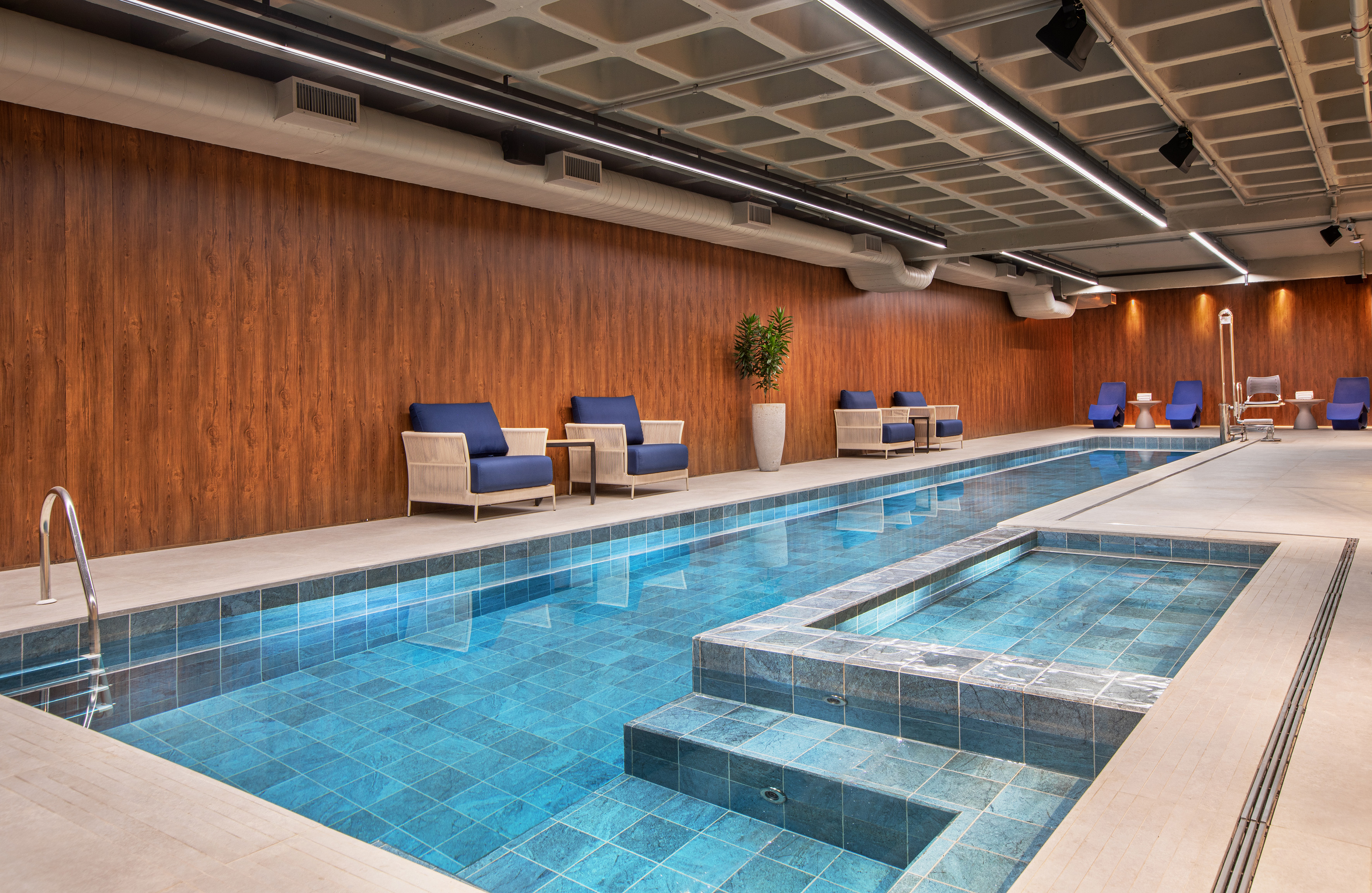 Modern indoor pool with blue tiles, lounge chairs, and wood paneling.