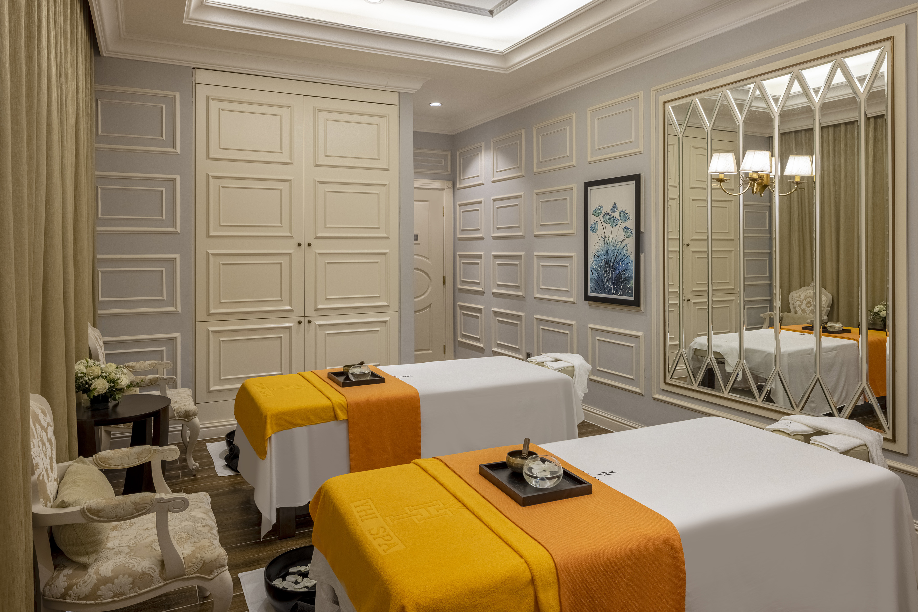 Elegant spa treatment room with two massage beds, orange throws, and patterned walls.