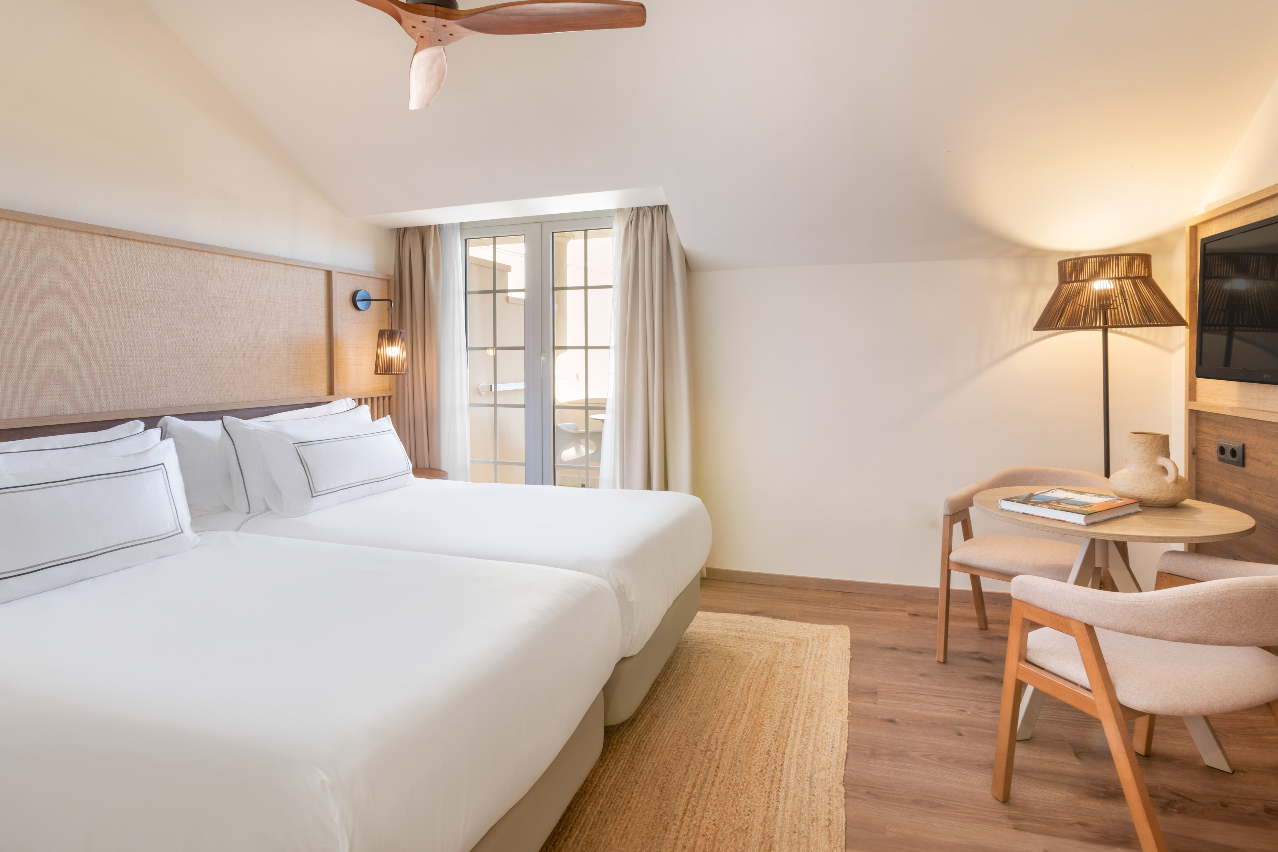 Cozy hotel room with twin beds, ceiling fan, balcony doors, and reading nook.