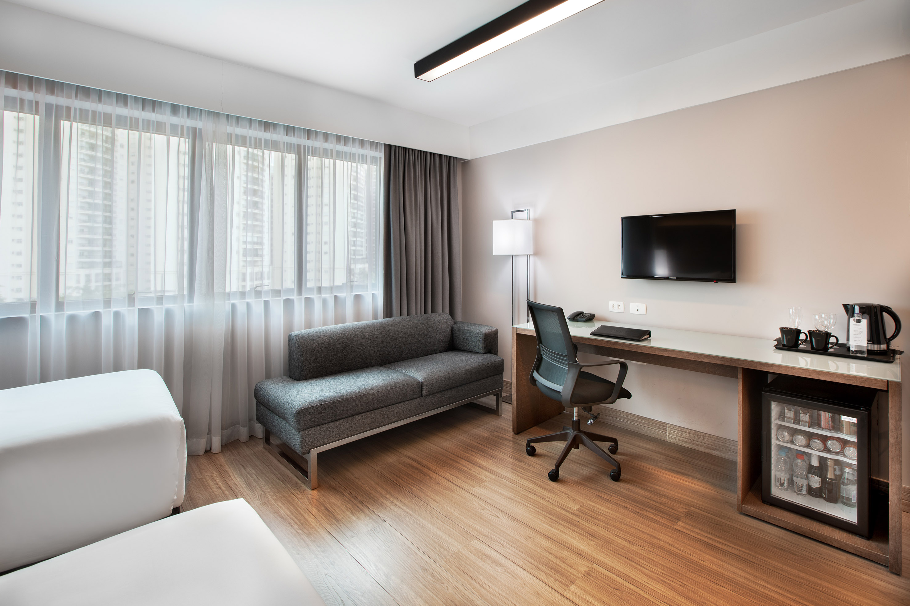 Modern hotel room with desk, TV, sofa, minibar, and large window.