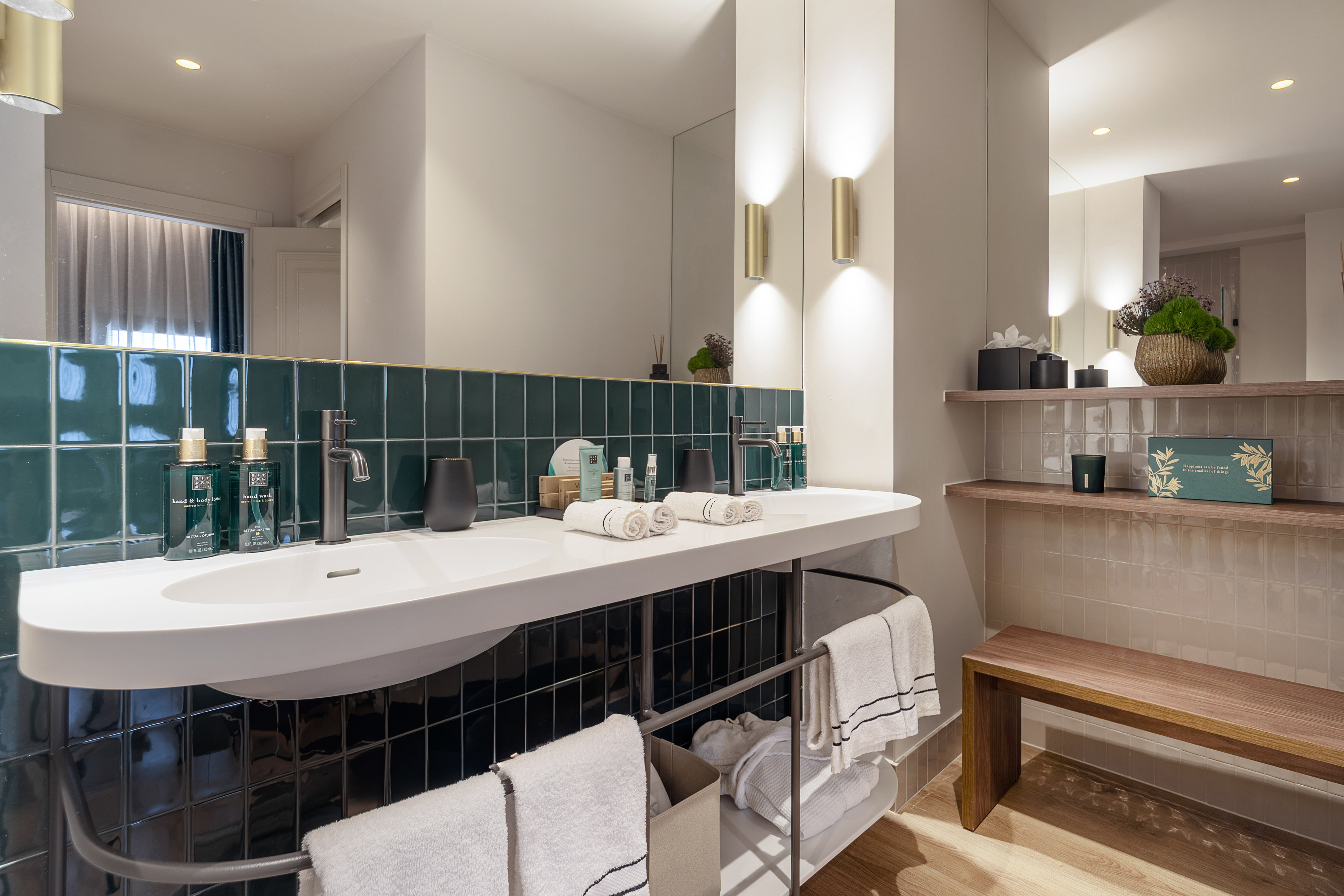 Stylish bathroom with dark green tiles, dual sinks, gold sconces, and wooden bench.