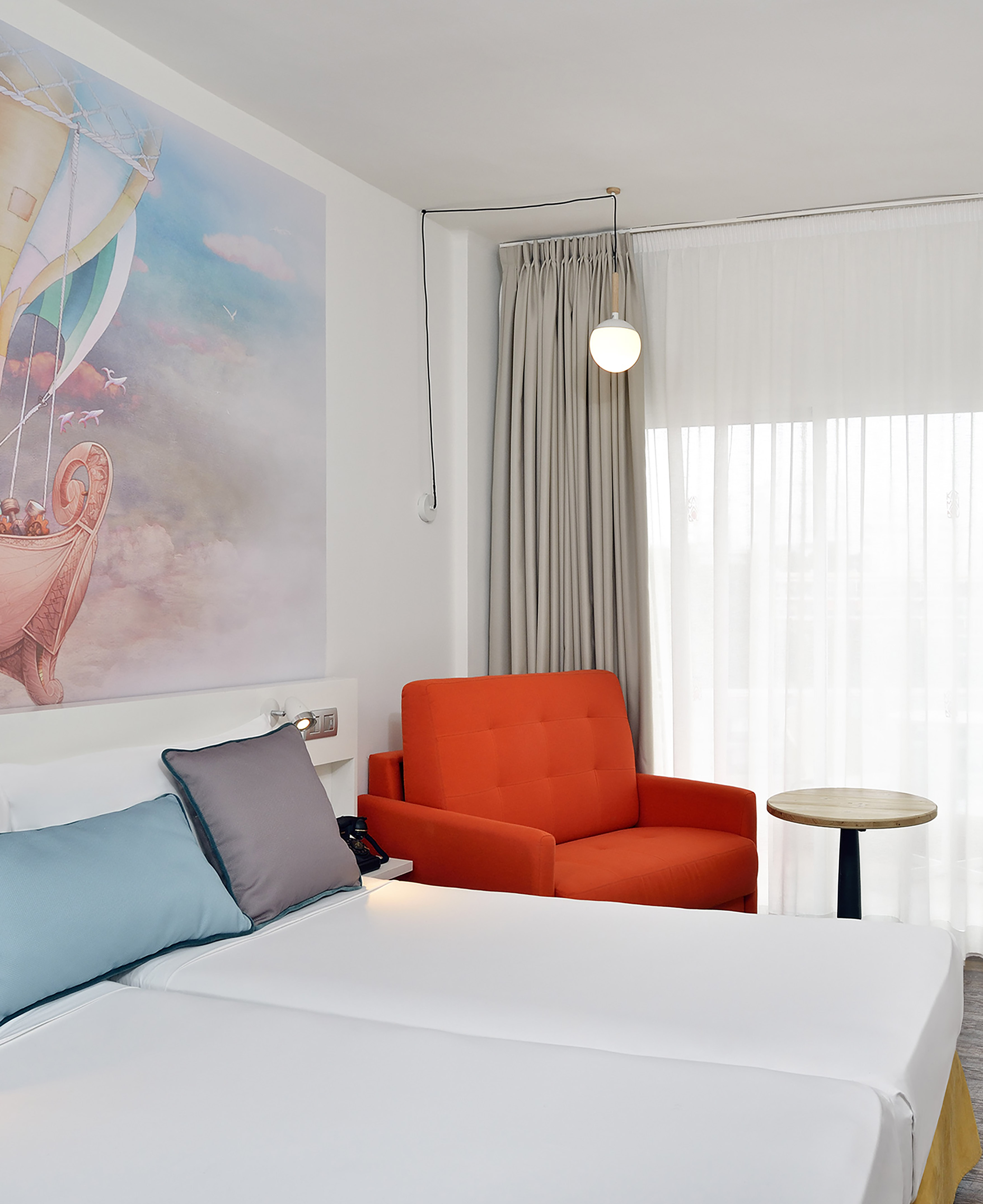 Bright hotel bedroom with hot air balloon mural, bed, orange armchair.