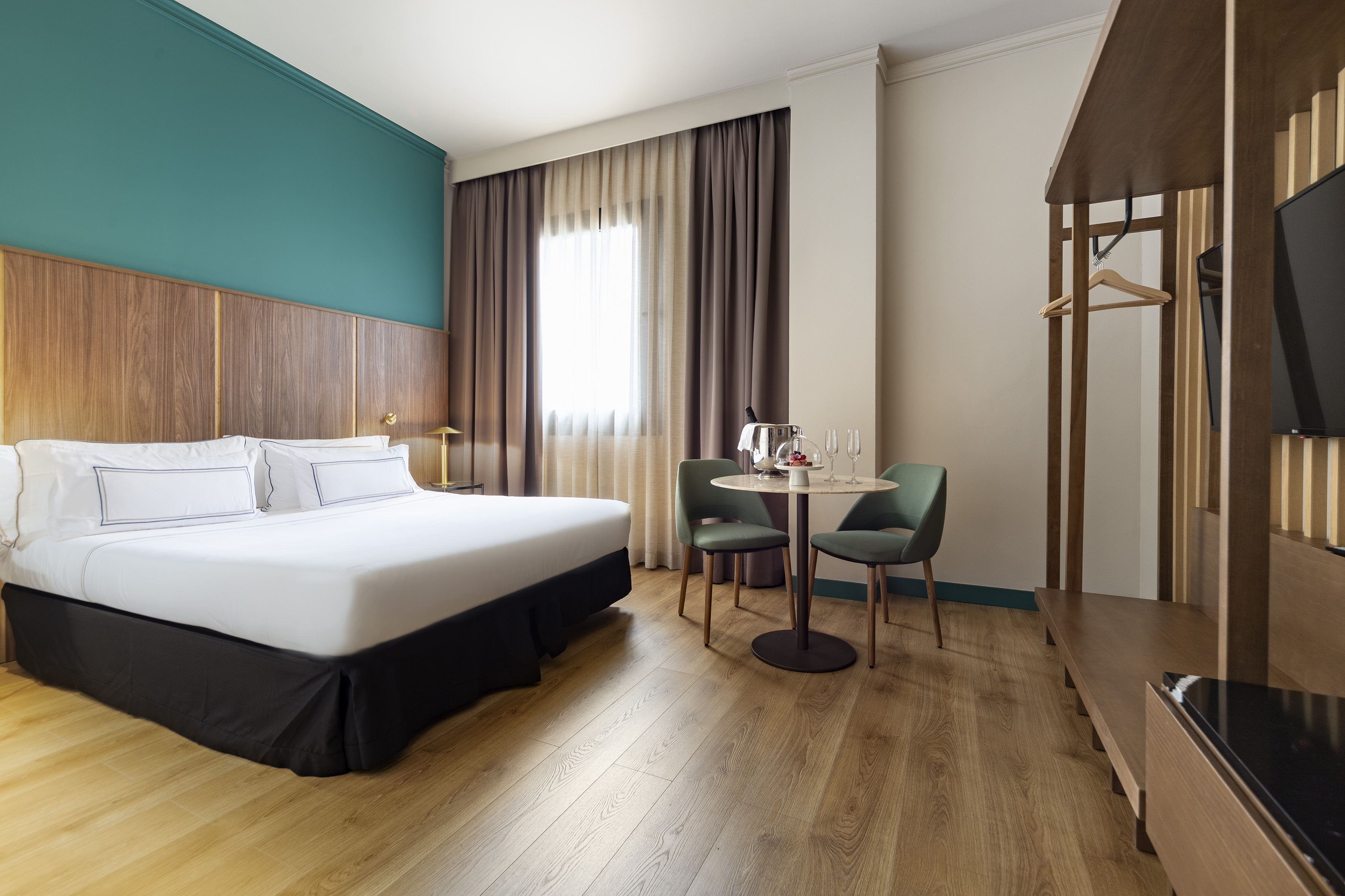 Modern hotel room: bed, teal accent wall, wood floor, and small sitting area.