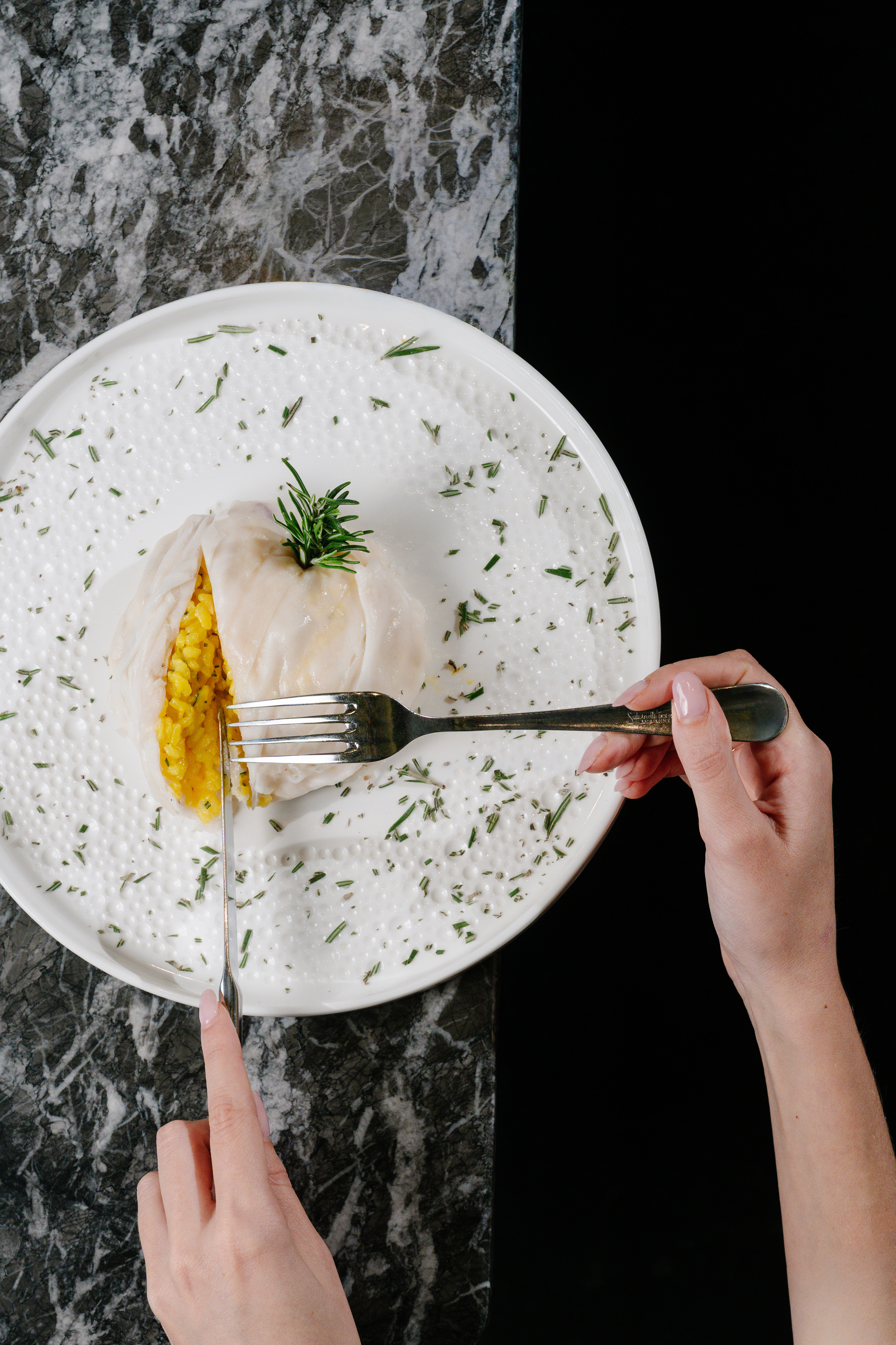Hands cutting a savory stuffed roll with yellow rice and rosemary on a white plate.
