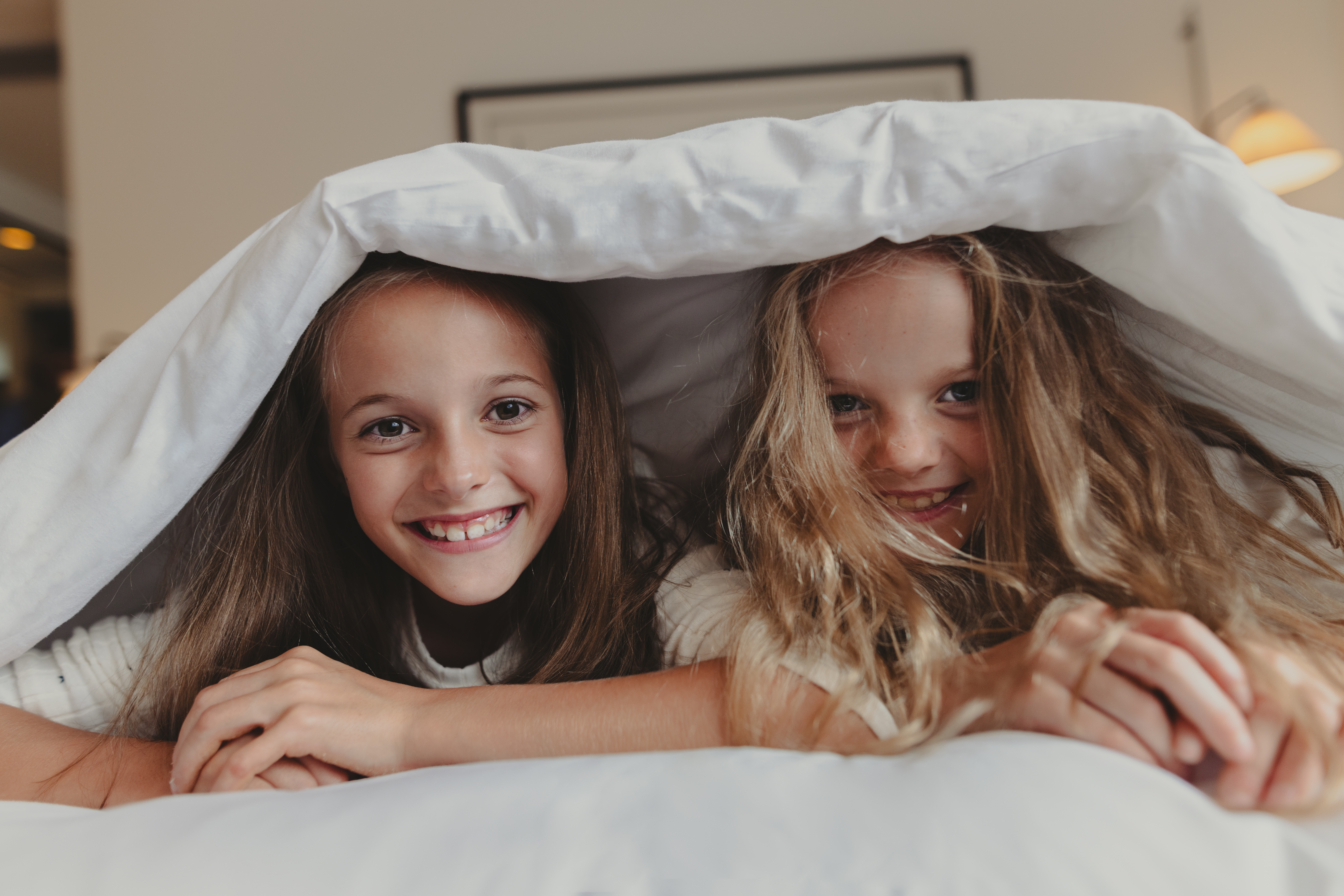 Two smiling girls peeking from under a white bed duvet.