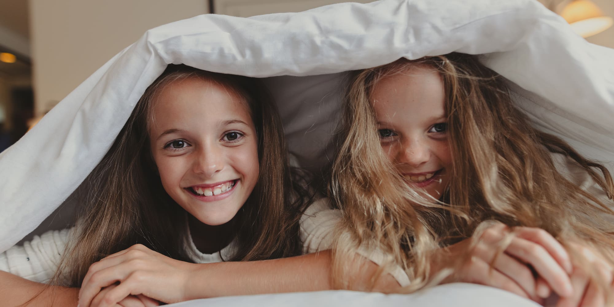 Two smiling girls peeking from under a white bed duvet.
