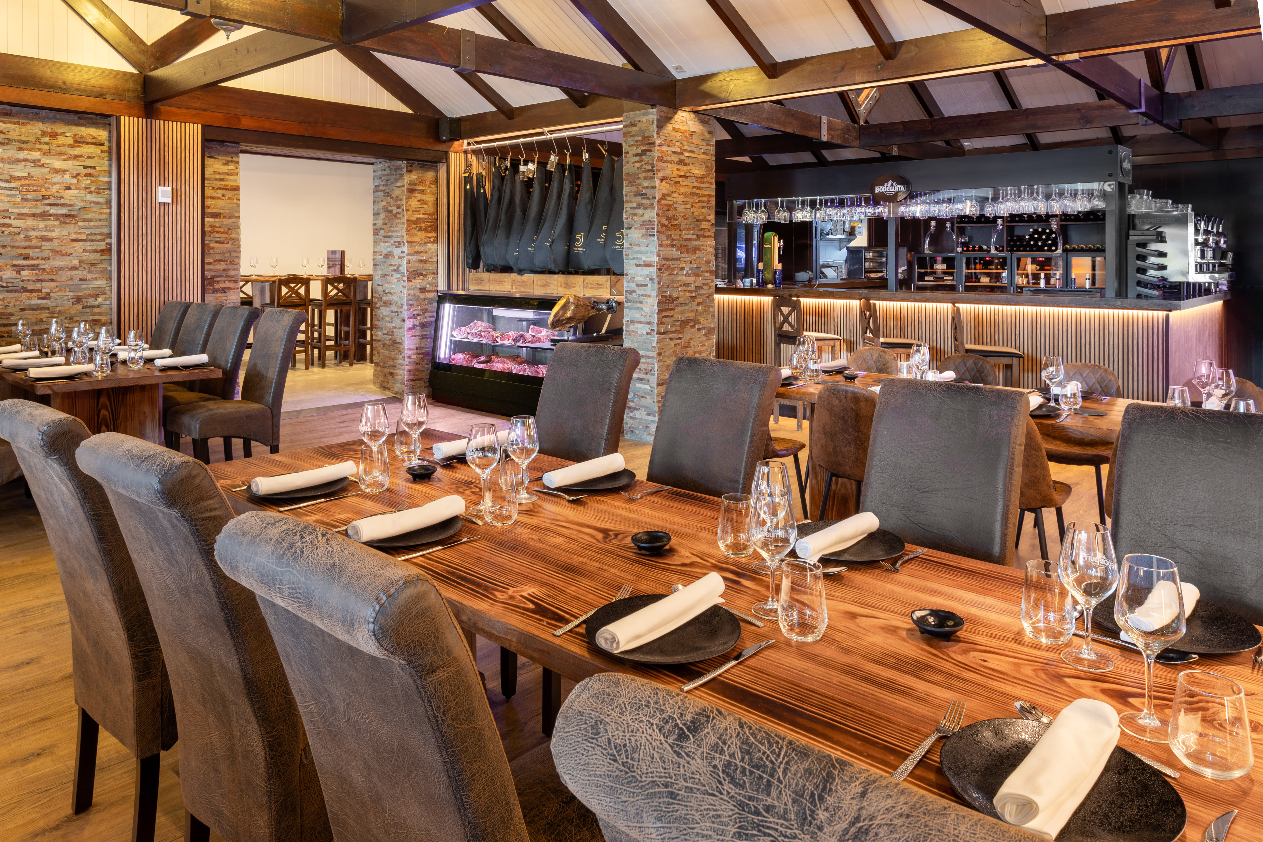 Upscale steakhouse dining room with set wooden tables, comfortable chairs, bar, and meat display.