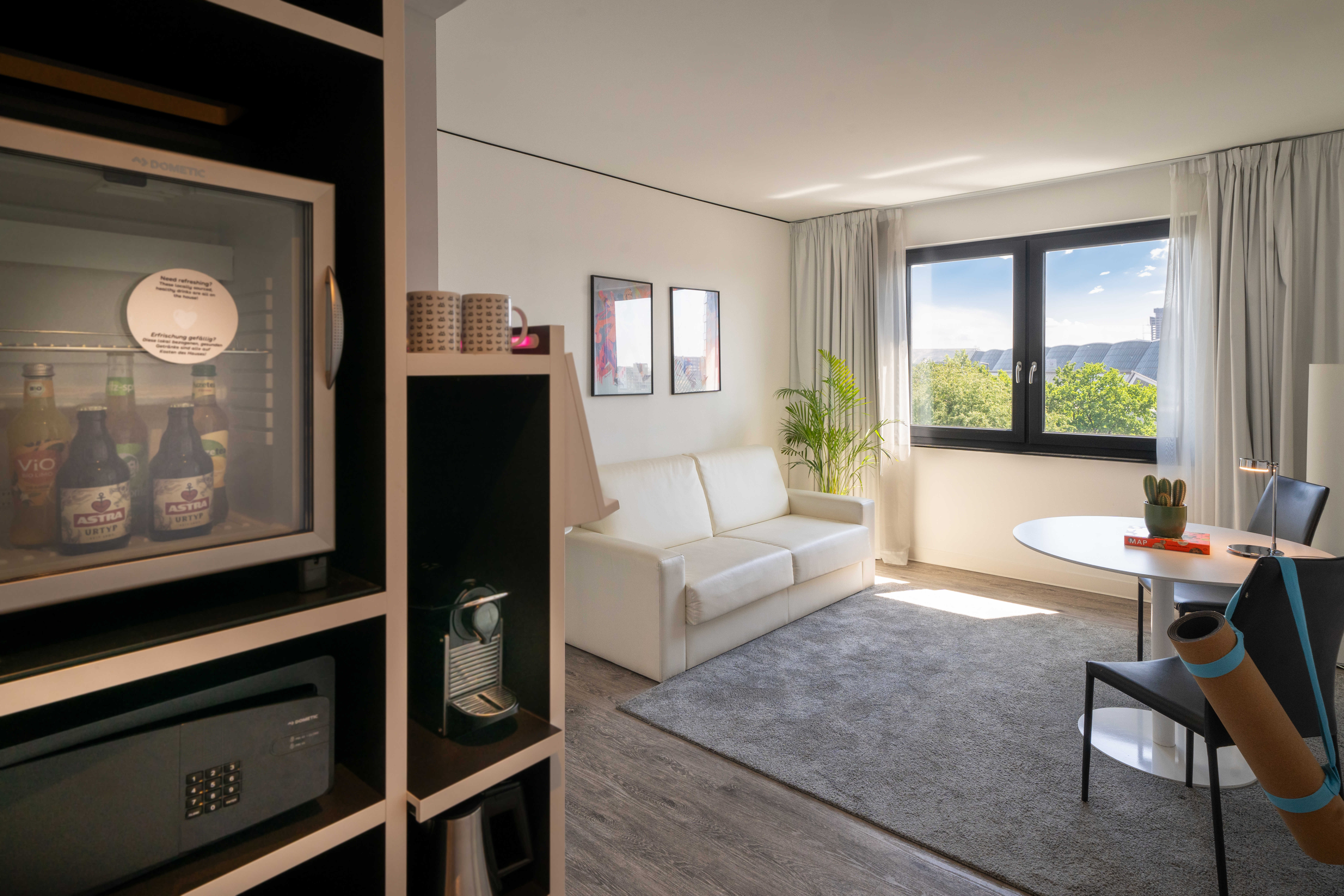 Bright hotel room with white sofa, minibar, coffee maker, and large window view.
