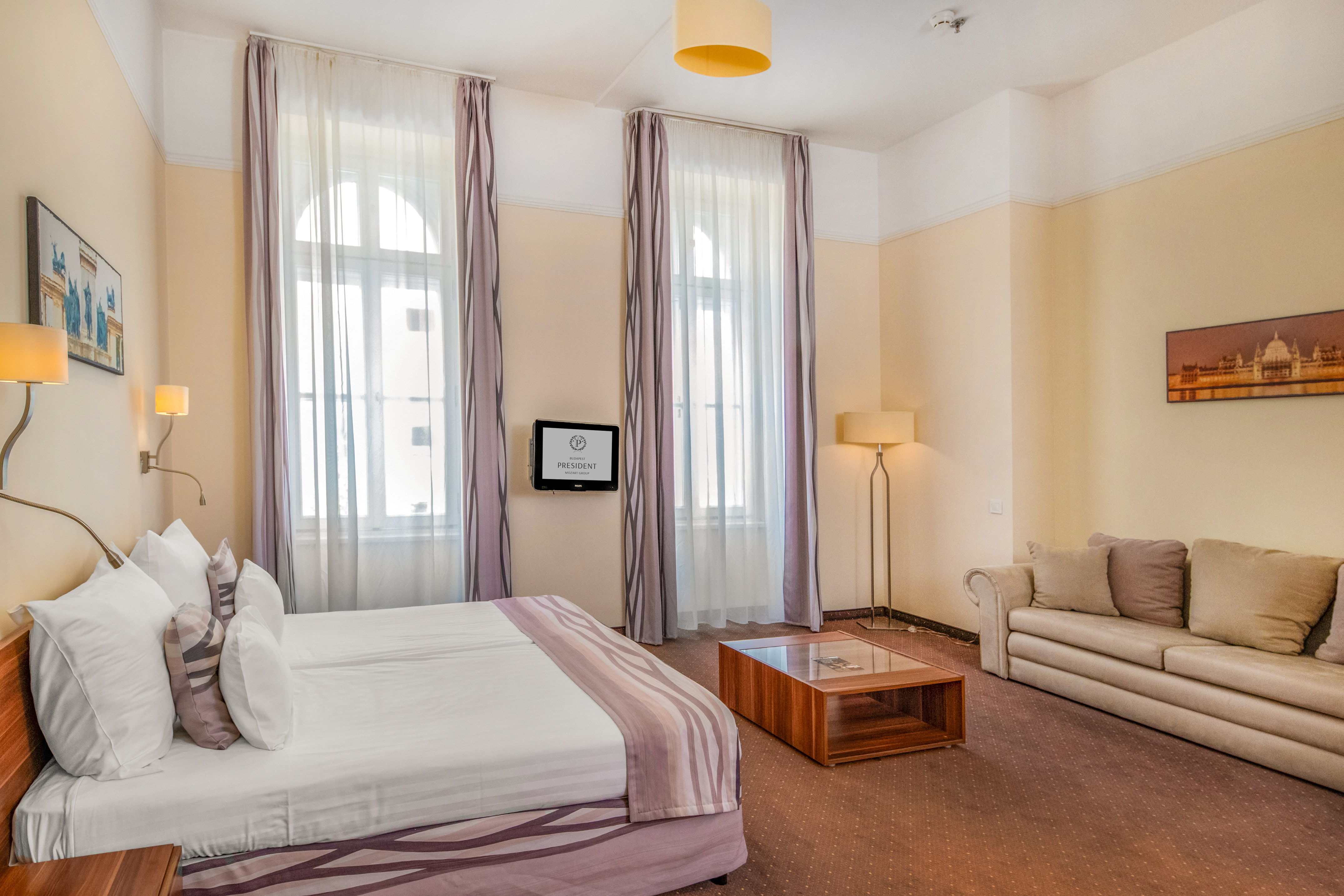 Spacious hotel room with twin beds, sofa, wall-mounted TV, and arched windows. - Junior Suite Classic - Hotel President Budapest Affiliated by Meliá, Budapest