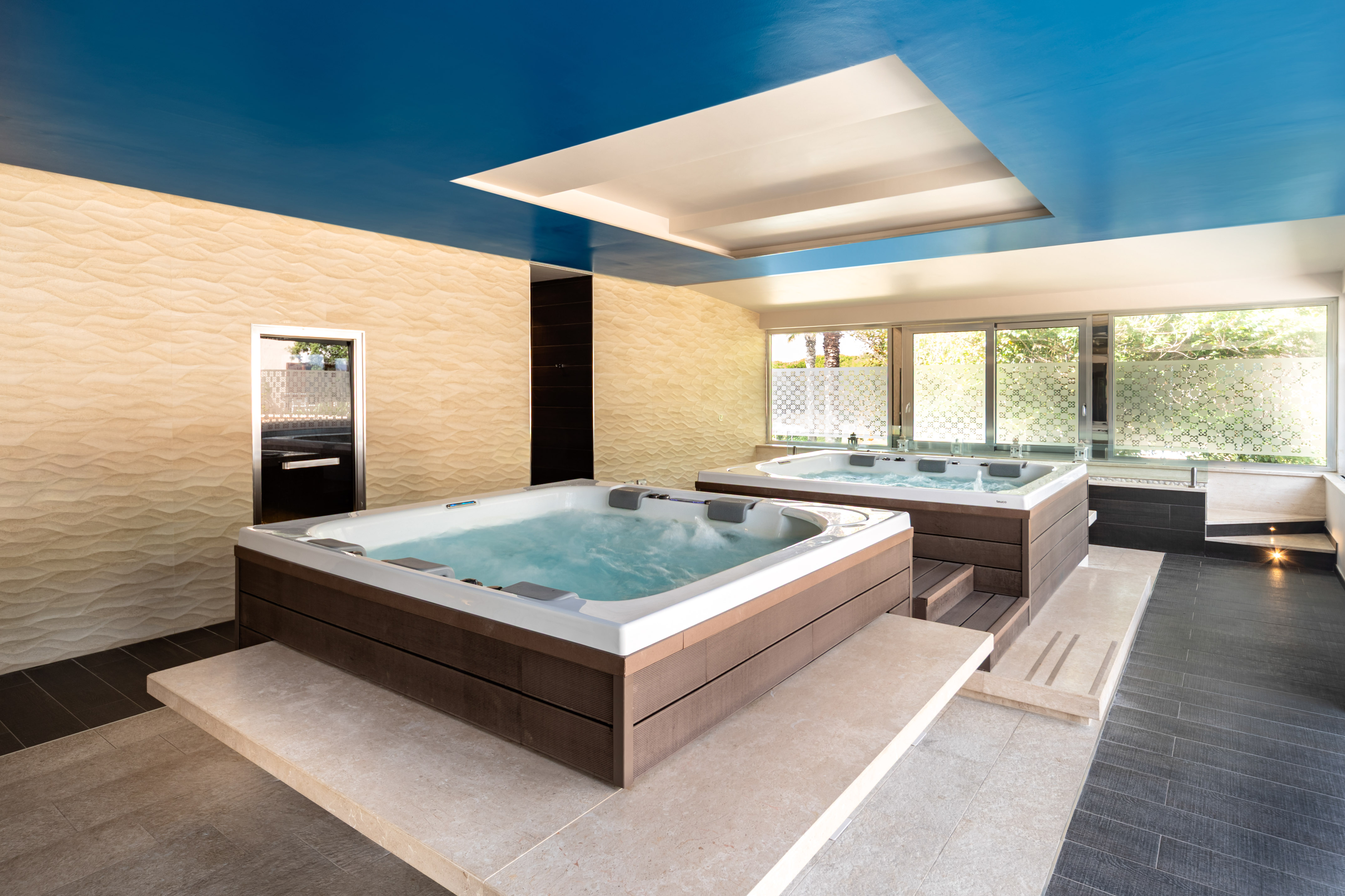 Two bubbling hot tubs in modern indoor spa room, blue ceiling, textured walls.
