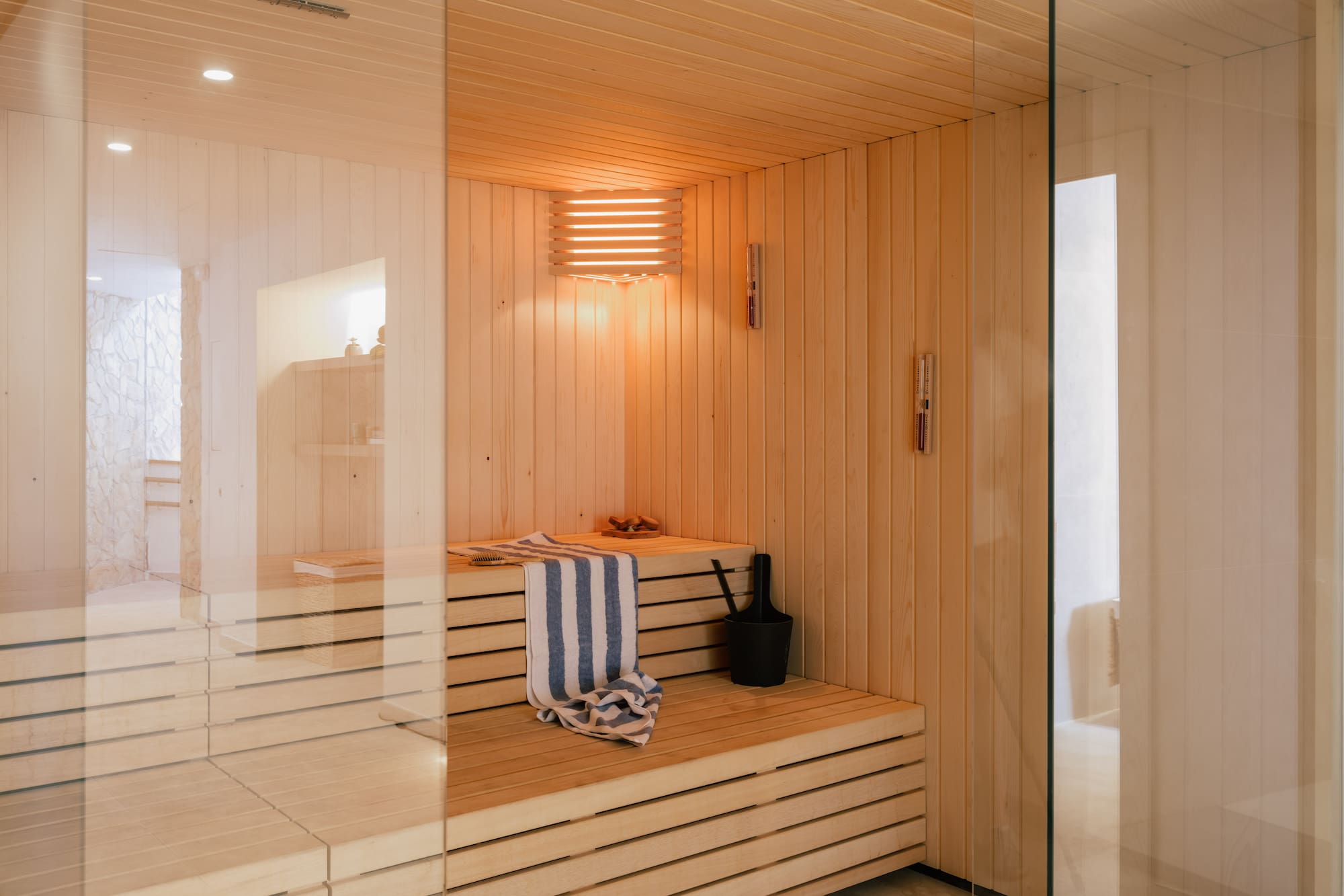Warm-lit wooden sauna with glass wall, slatted benches, towel, and bucket.