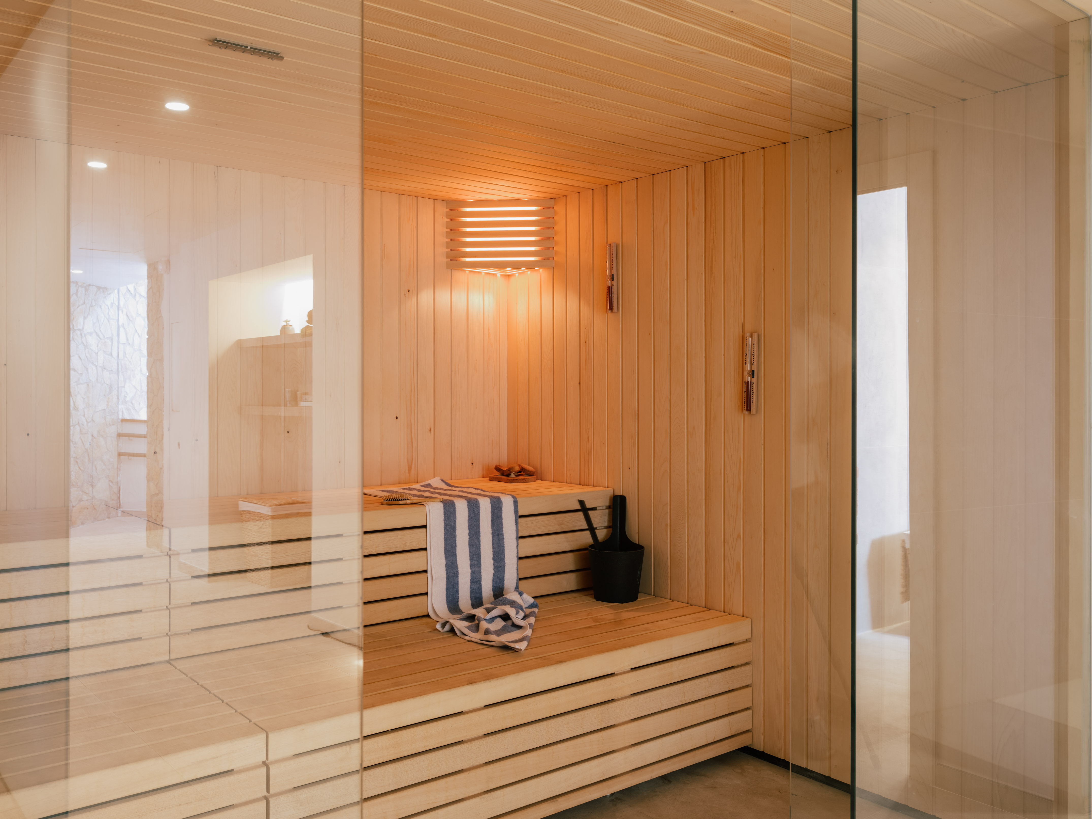 Warm-lit wooden sauna with glass wall, slatted benches, towel, and bucket.