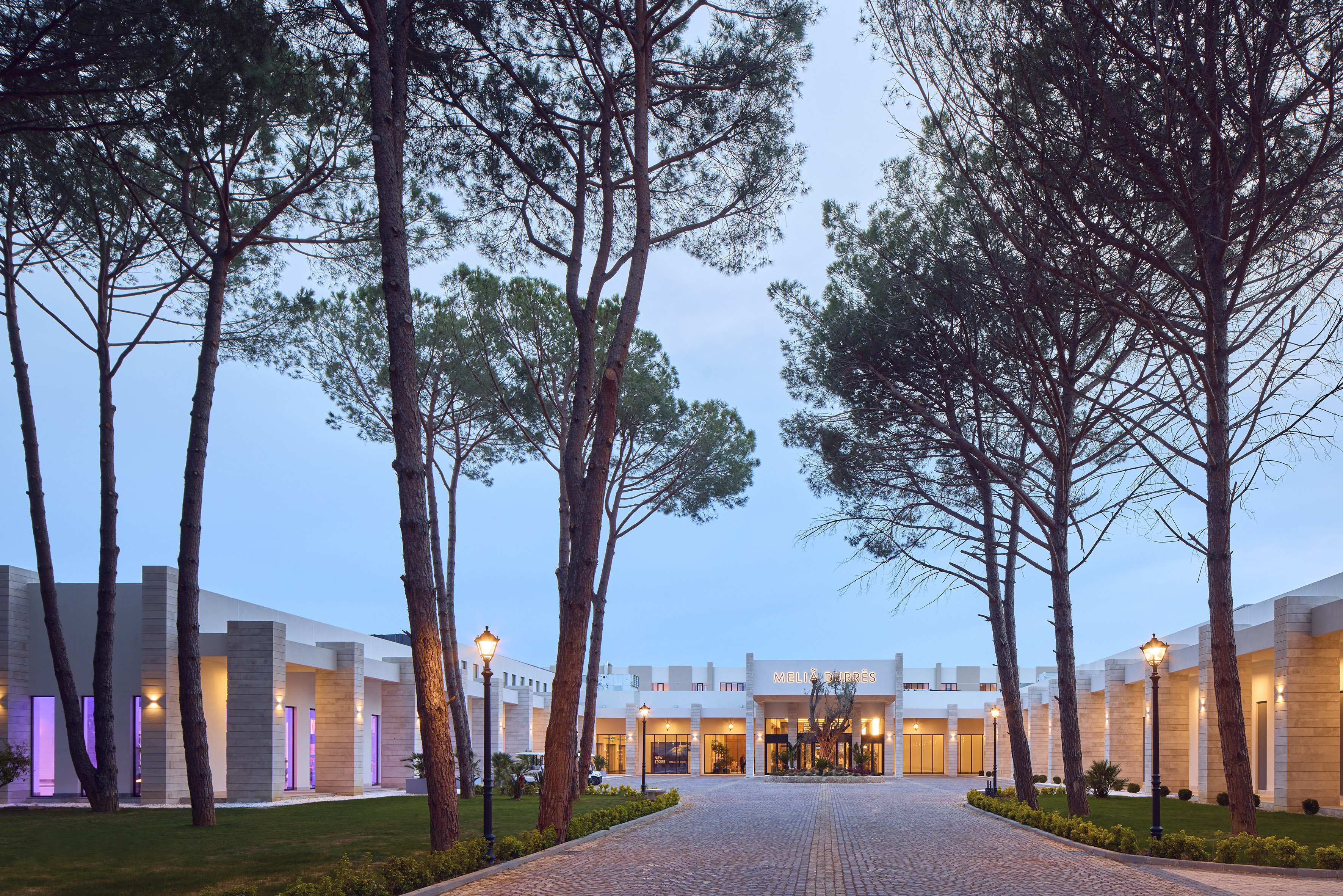 Modern Melia Durres hotel entrance, paved driveway, tall pine trees at dusk.