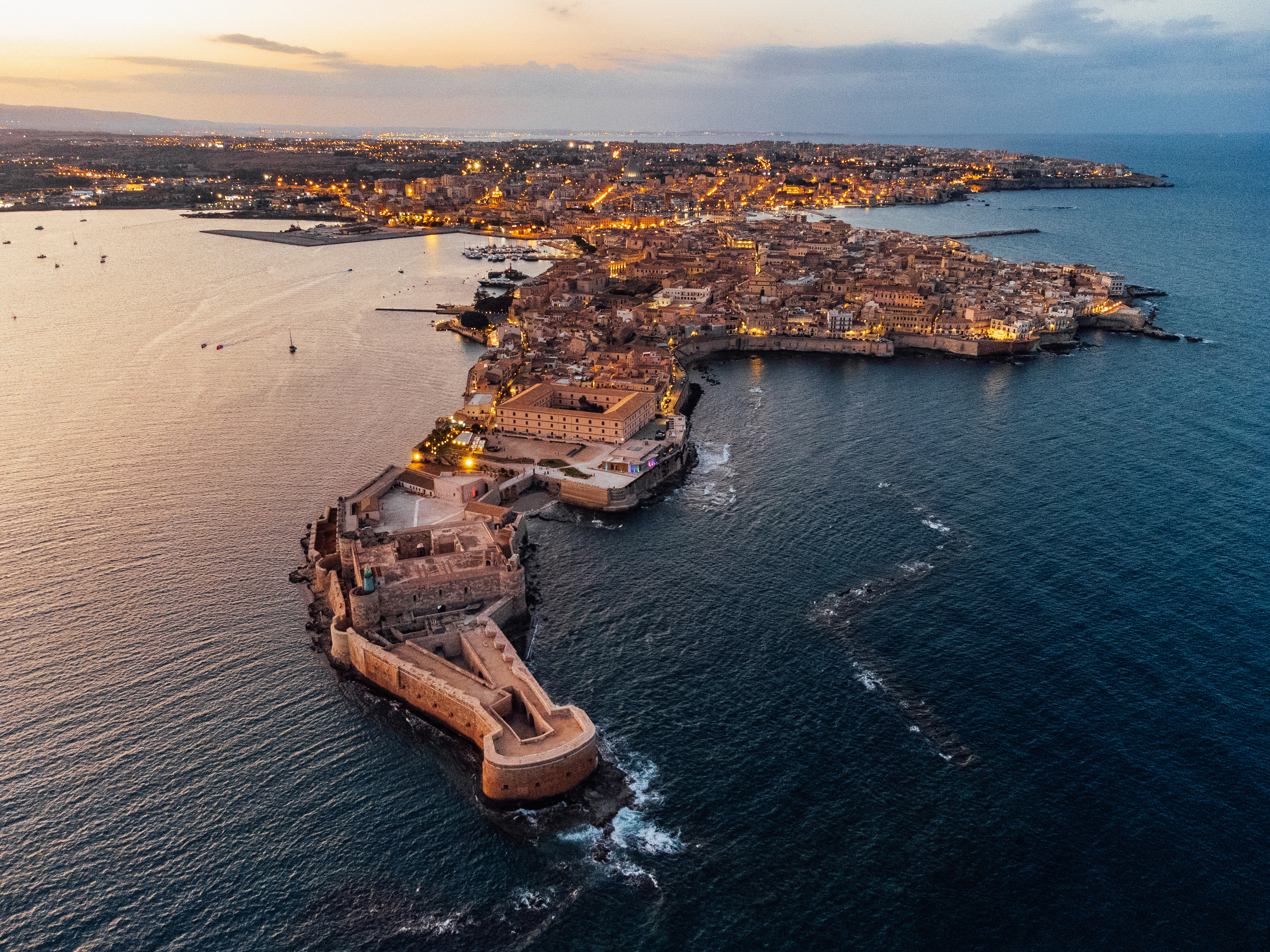 Vista aérea de Ortigia, Siracusa, con fortaleza y ciudad iluminada al anochecer.