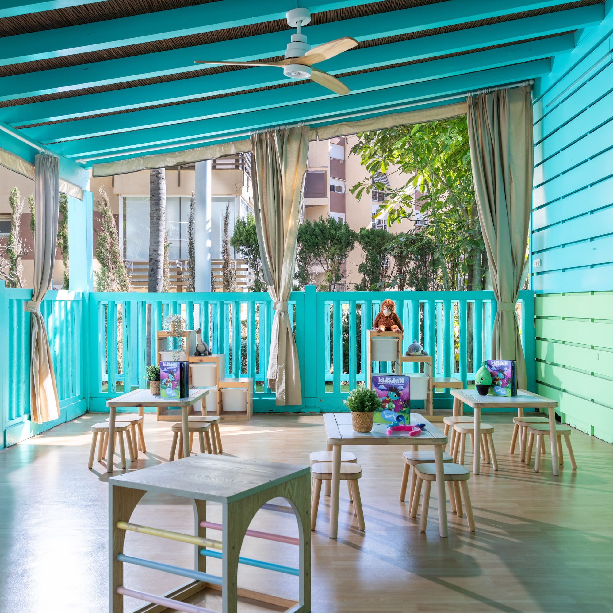 Bright turquoise kids club outdoor play area with small wooden tables and stools.