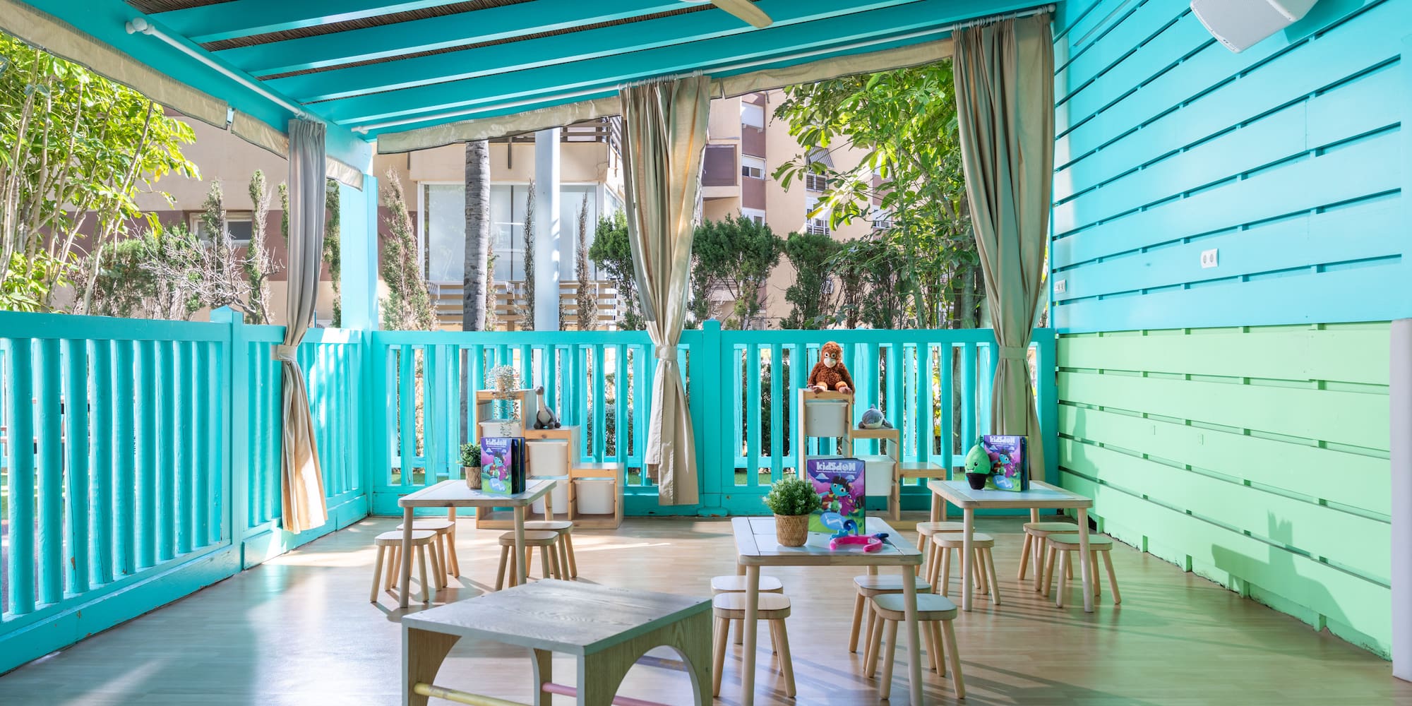 Bright turquoise kids club outdoor play area with small wooden tables and stools.