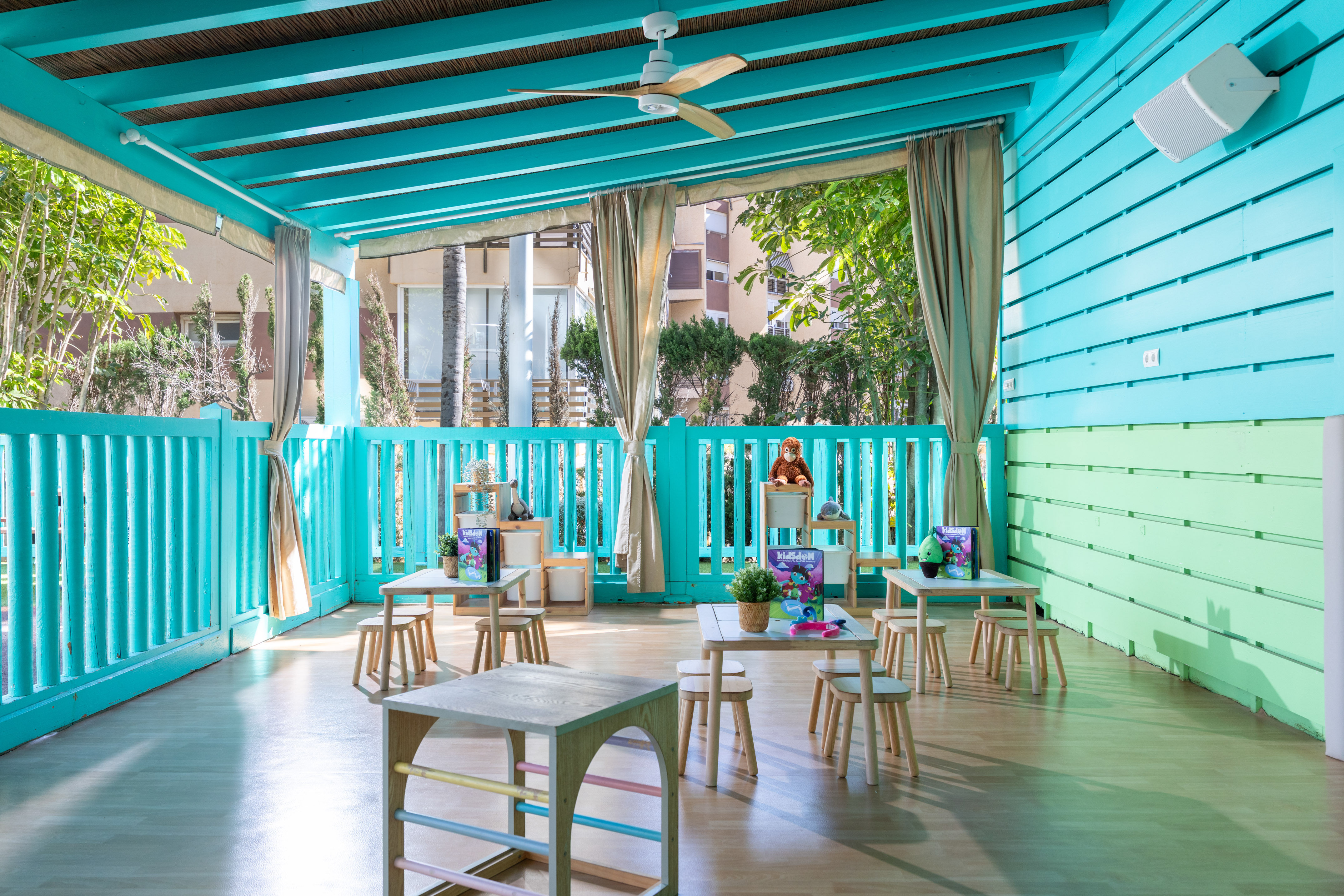 Bright turquoise kids club outdoor play area with small wooden tables and stools.