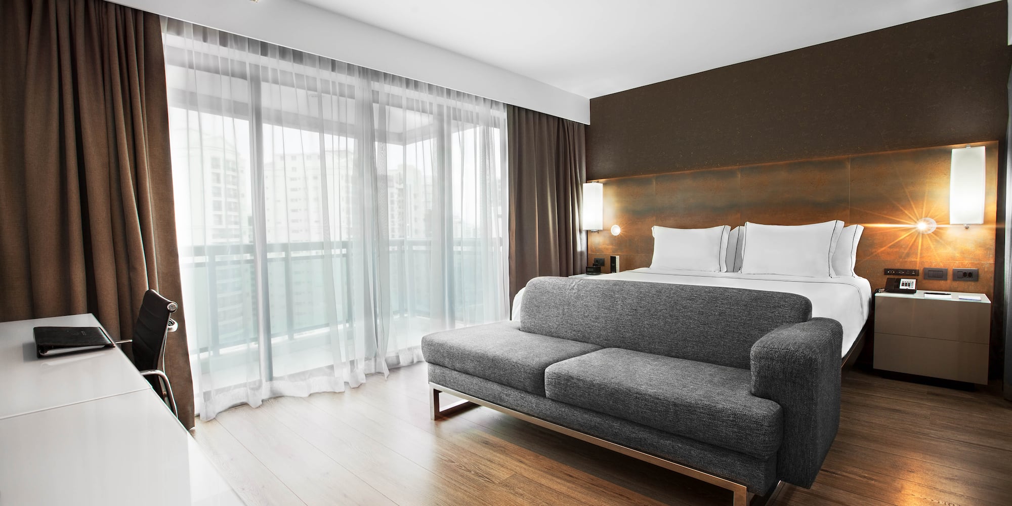 Luxury hotel suite with modern king bed, grey sofa, desk, and balcony.