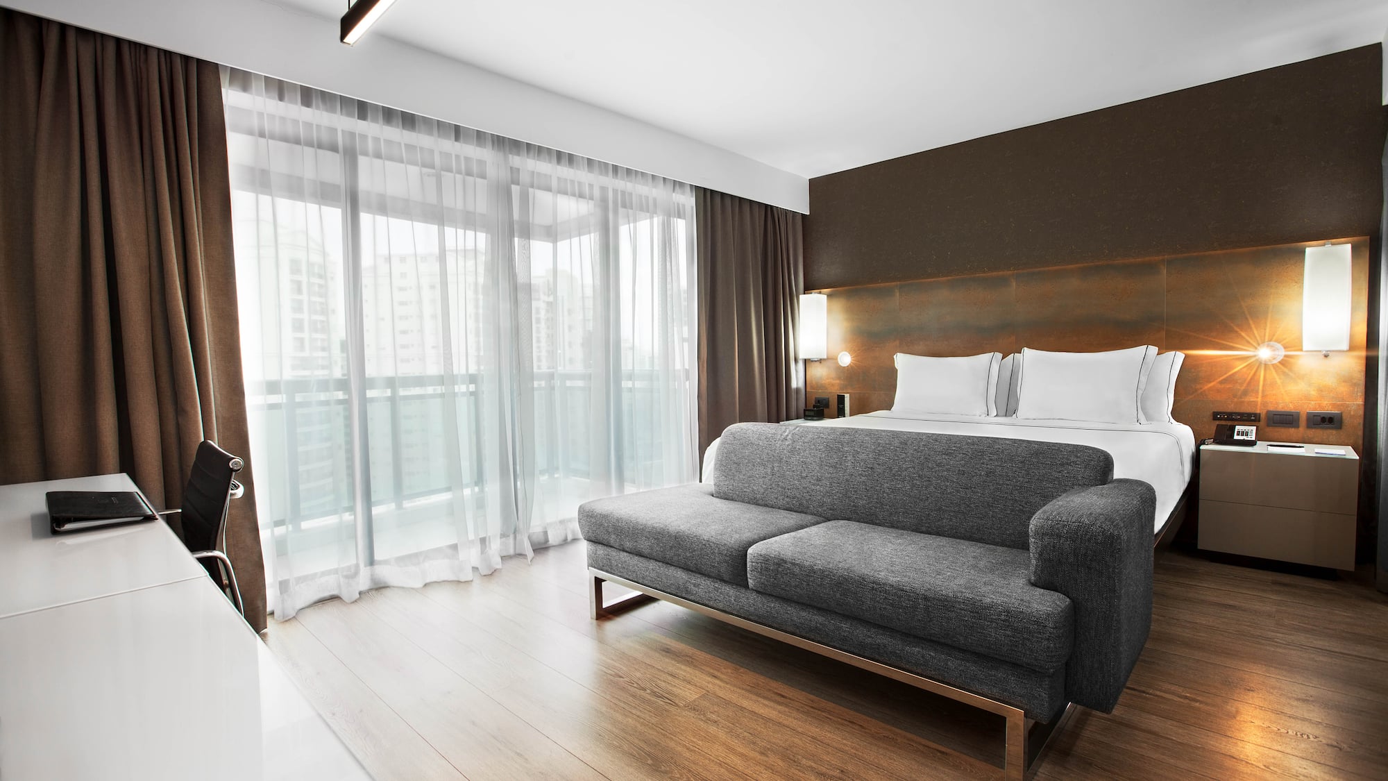 Modern hotel room with bed, grey sofa, desk, and large balcony windows.