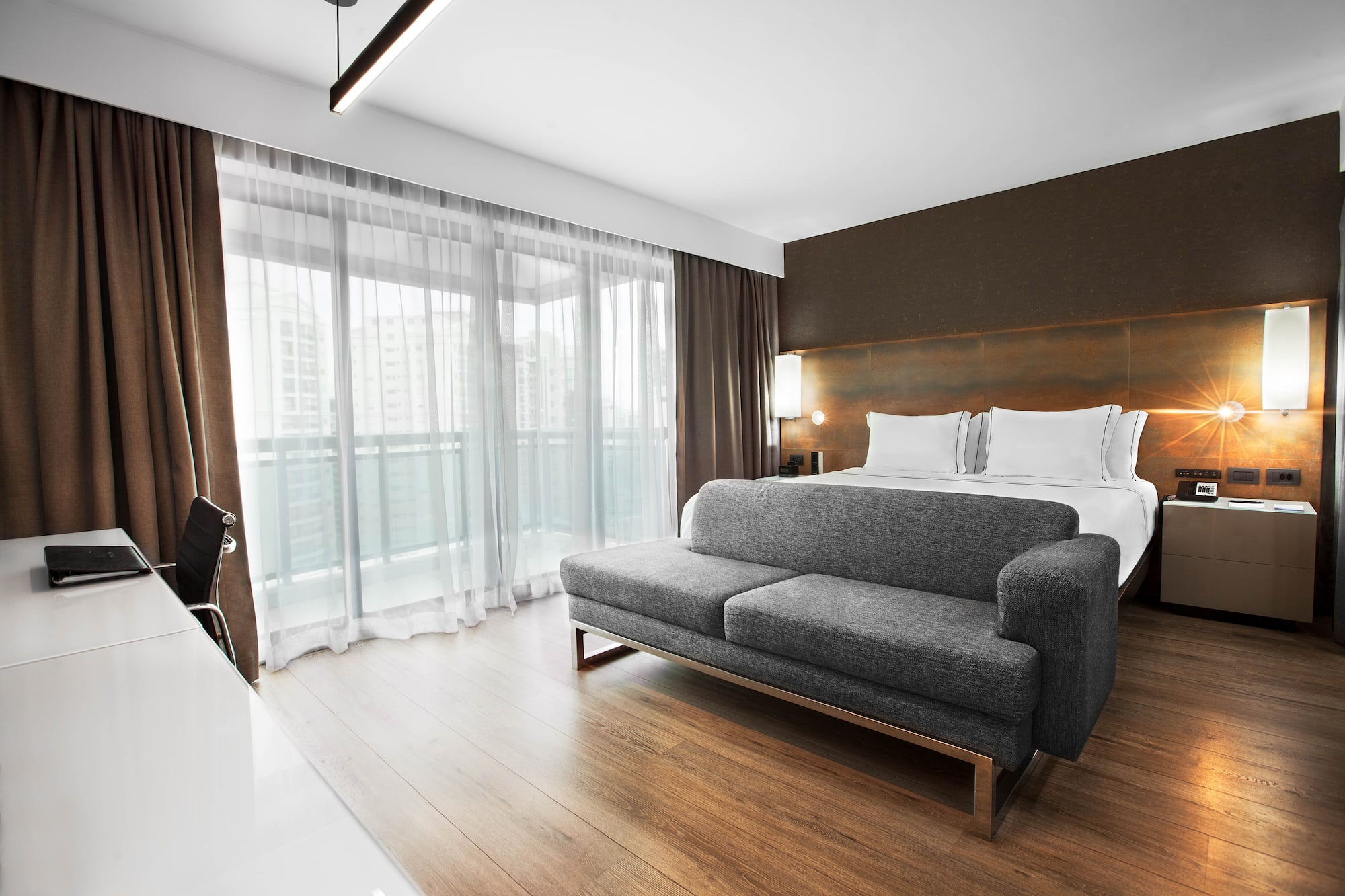Modern hotel room with bed, grey sofa, desk, and large balcony windows.