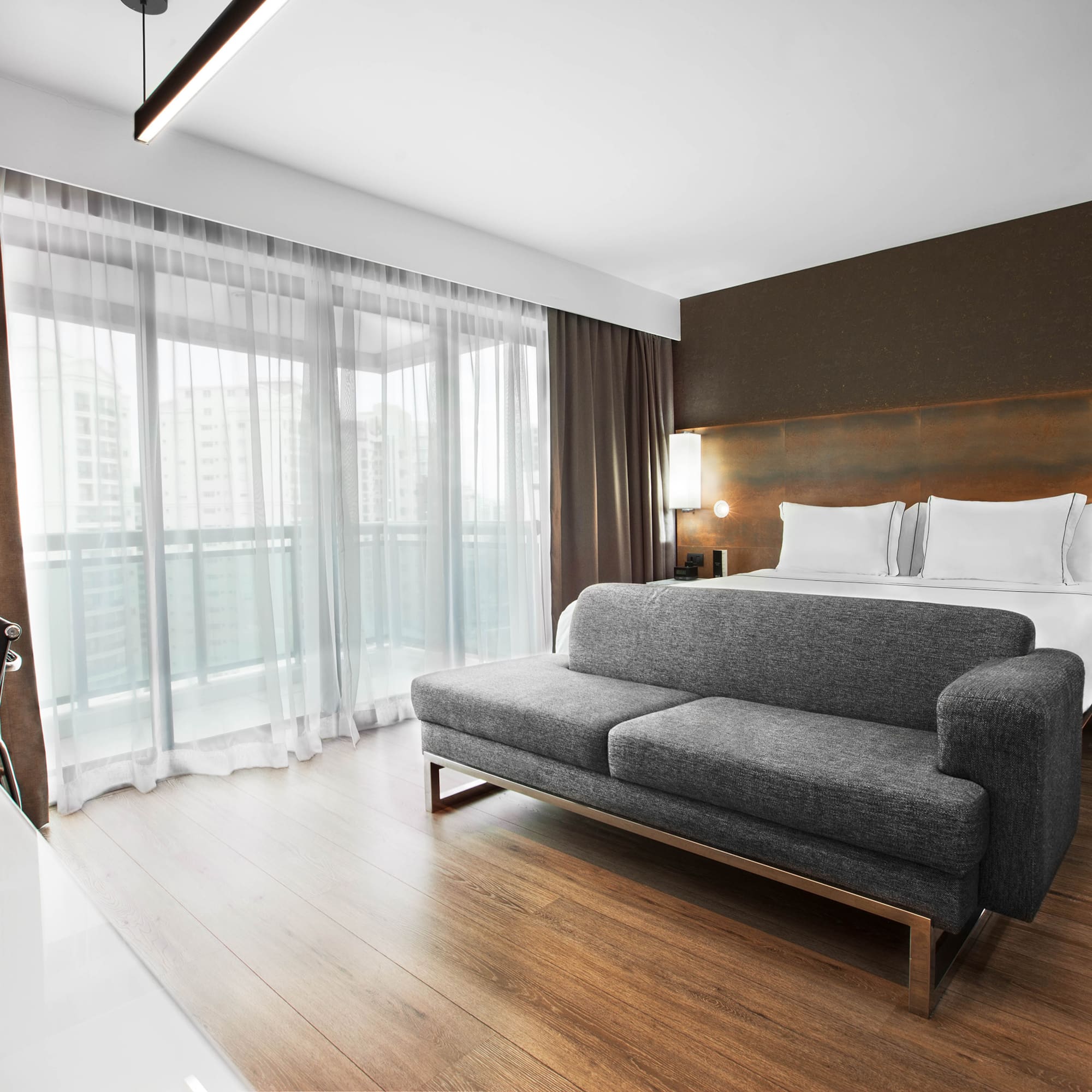 Modern hotel room with bed, grey sofa, desk, and large balcony windows.