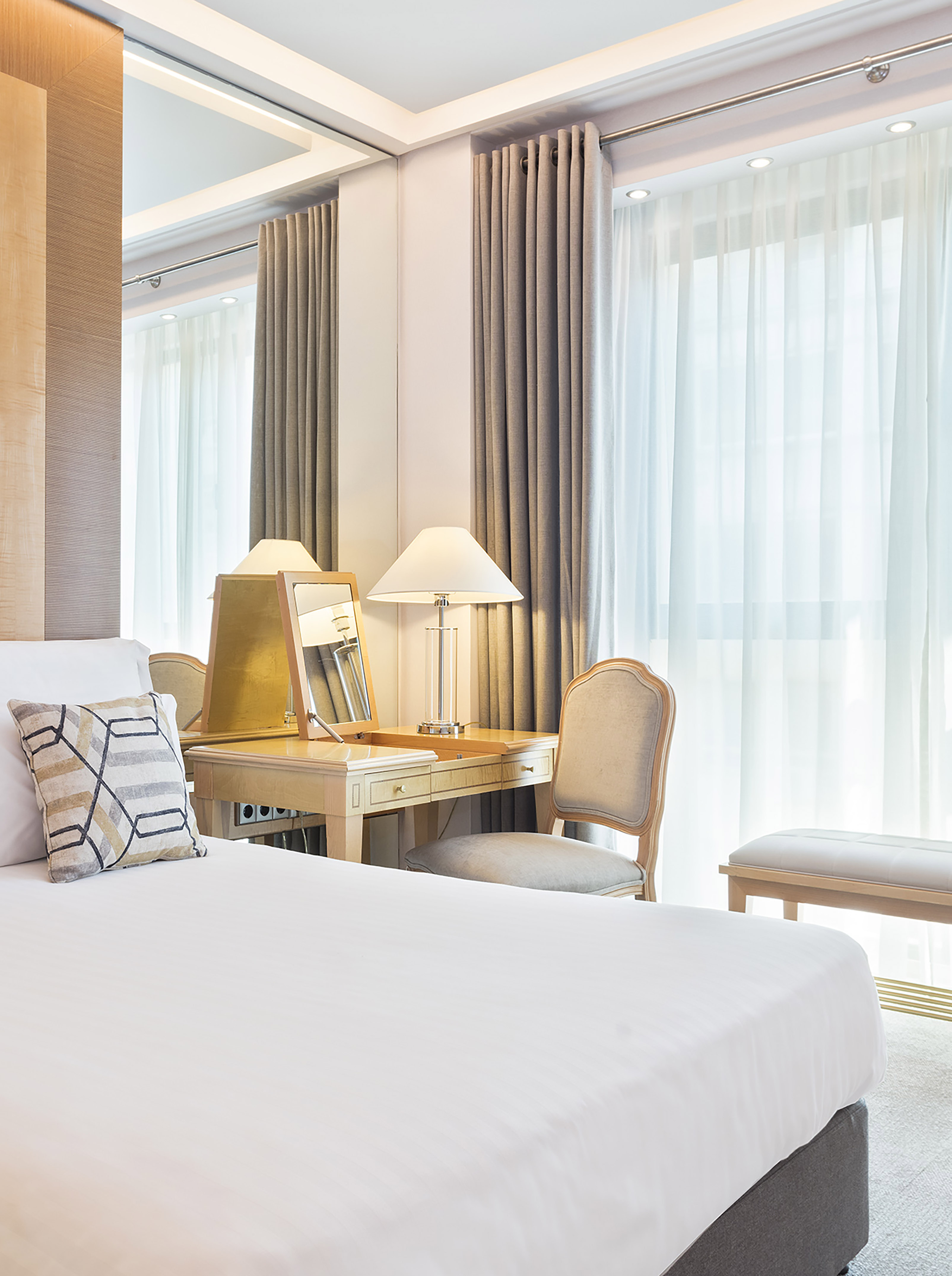 Hotel room with bed, dressing table, mirror, lamp, and sheer curtains.