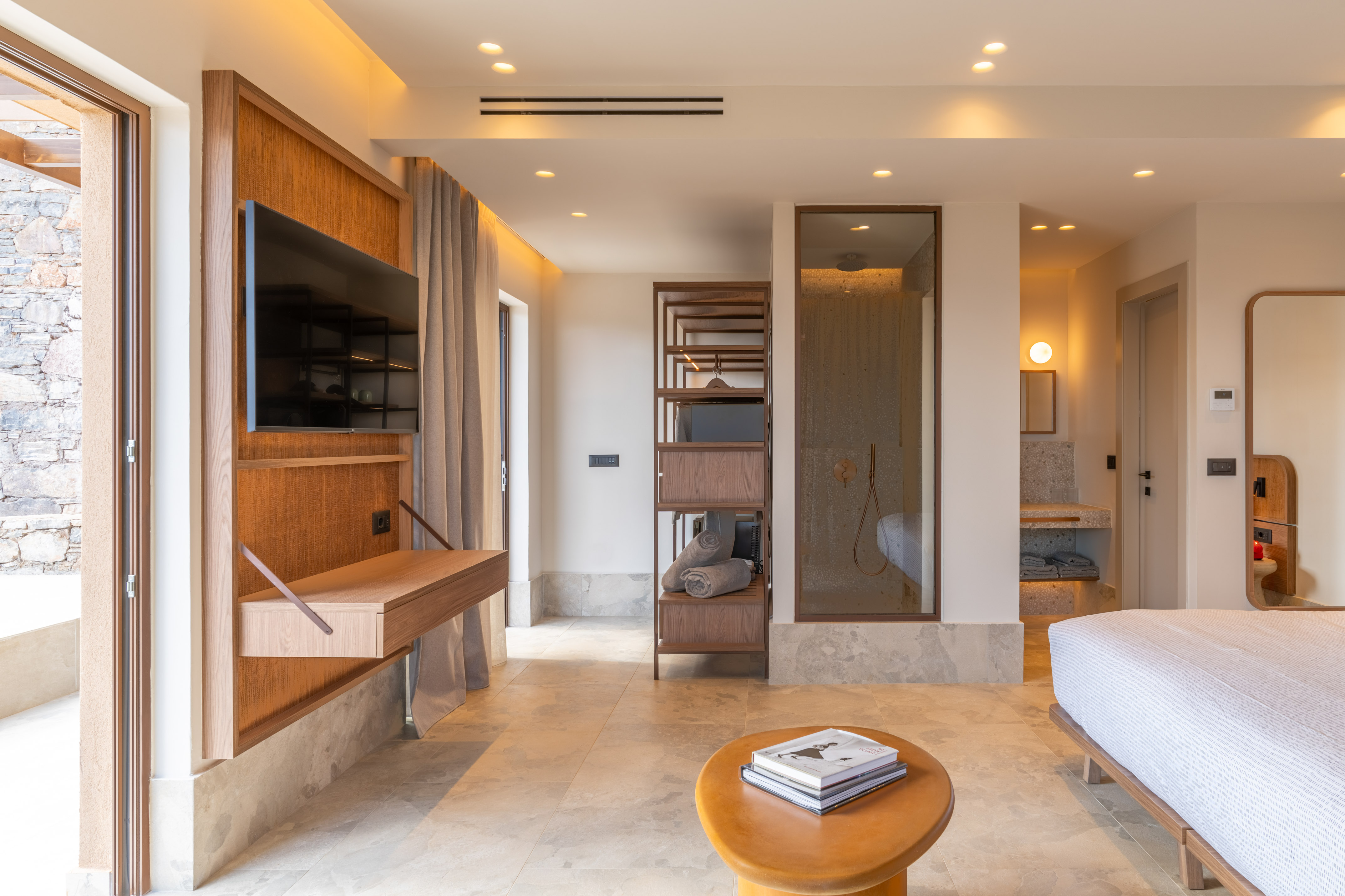 Modern luxury hotel room with wall TV, open glass shower, wood accents.