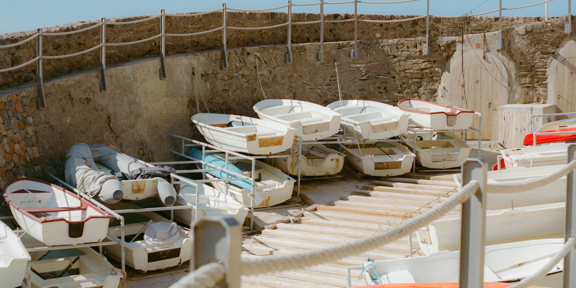White and colorful small boats stacked on dry storage racks by a stone wall.