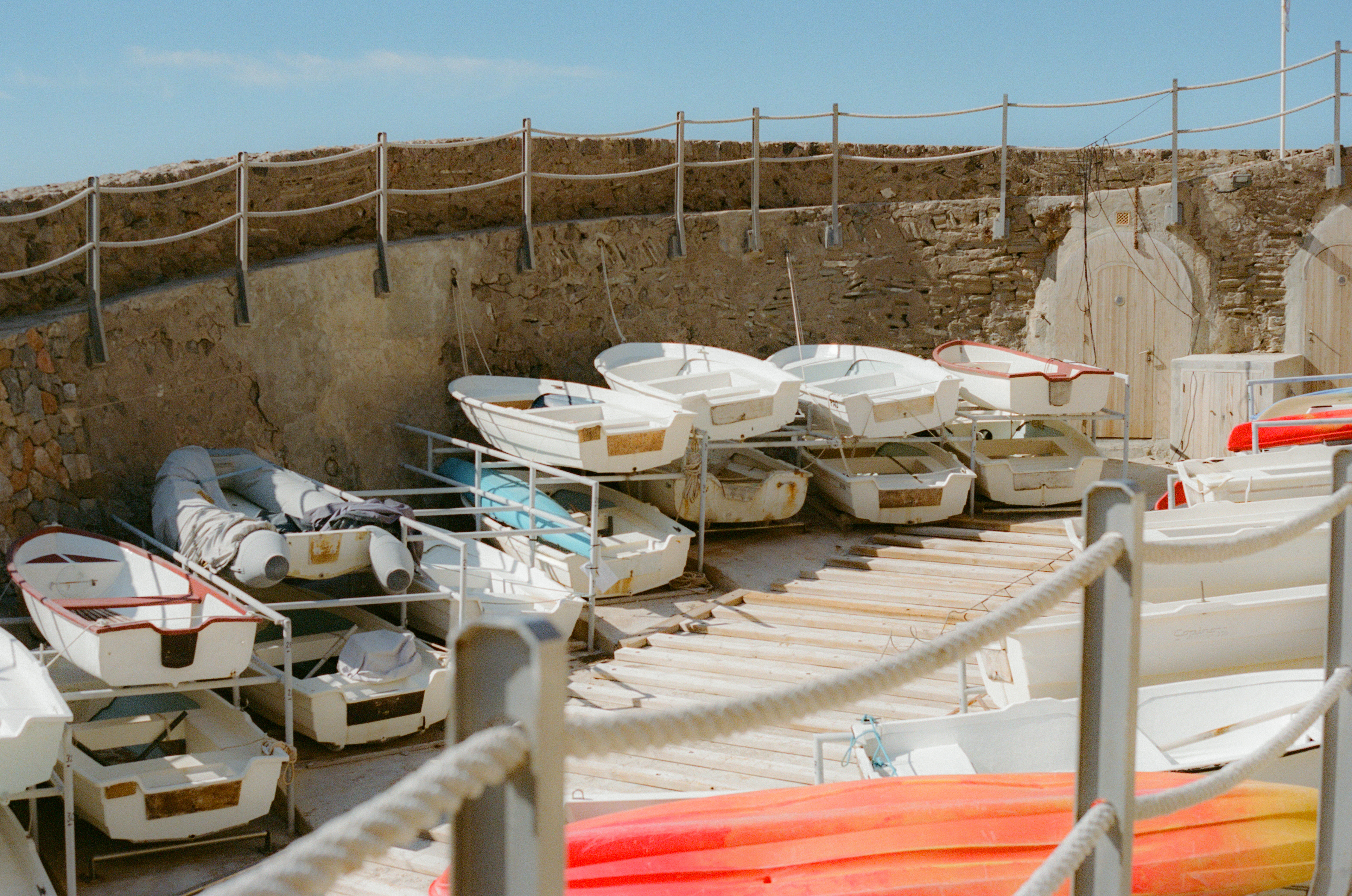 White and colorful small boats stacked on dry storage racks by a stone wall.