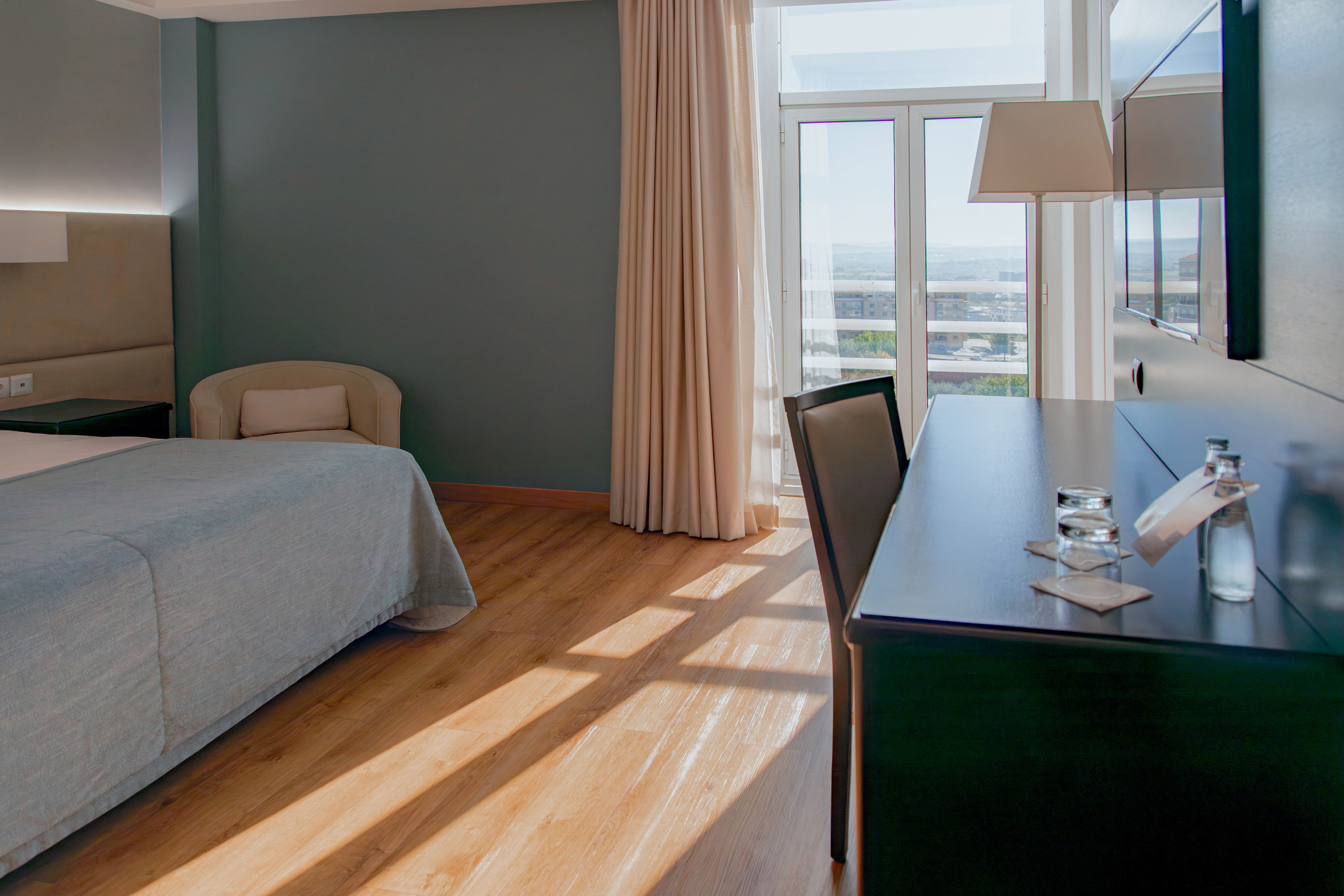 Spacious hotel bedroom with city view, desk, bed, and natural light on wood floor.