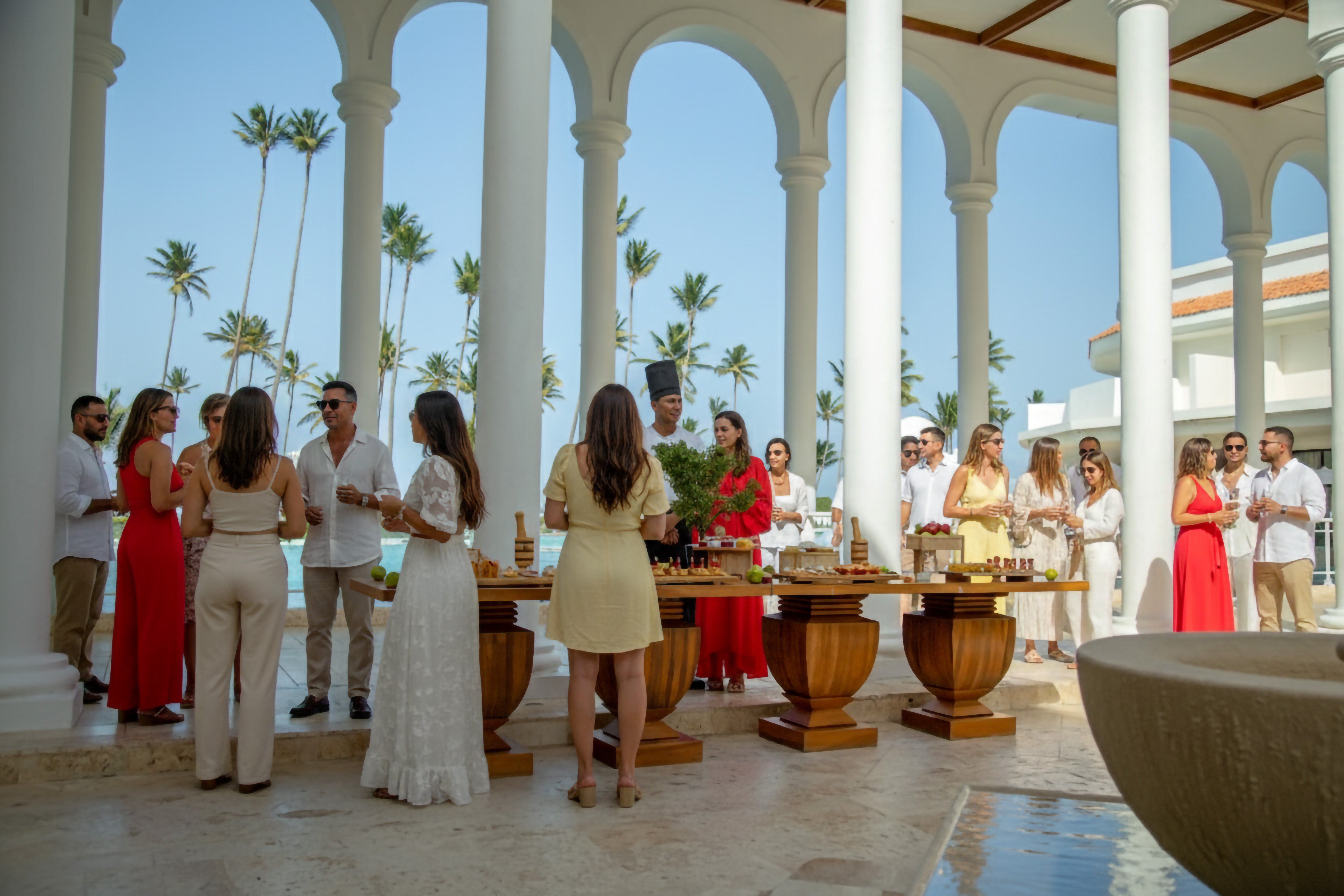 Guests mingle at an outdoor tropical resort event with a food table and palm trees.