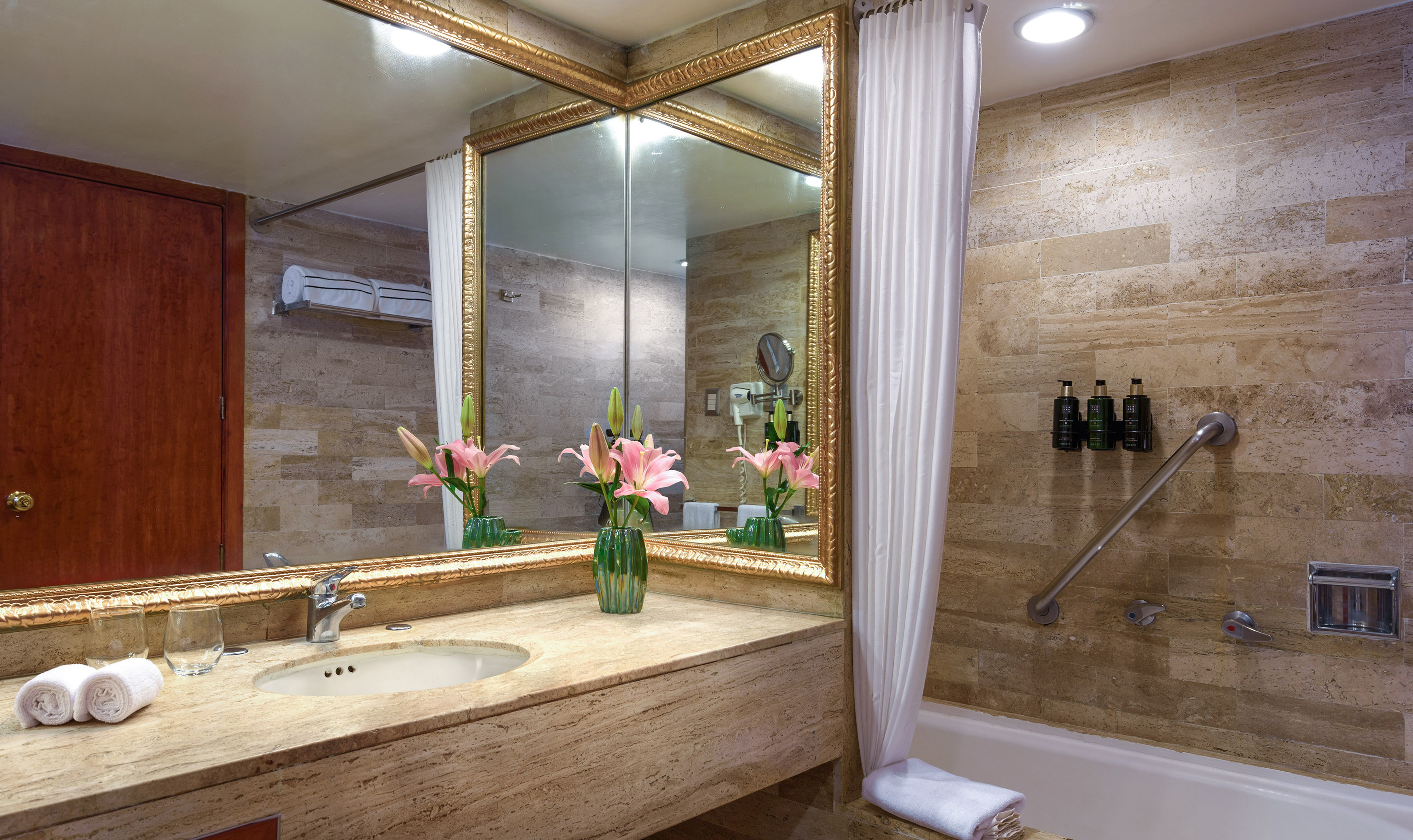 Elegant hotel bathroom with stone vanity, gold mirror, sink, and bathtub.
