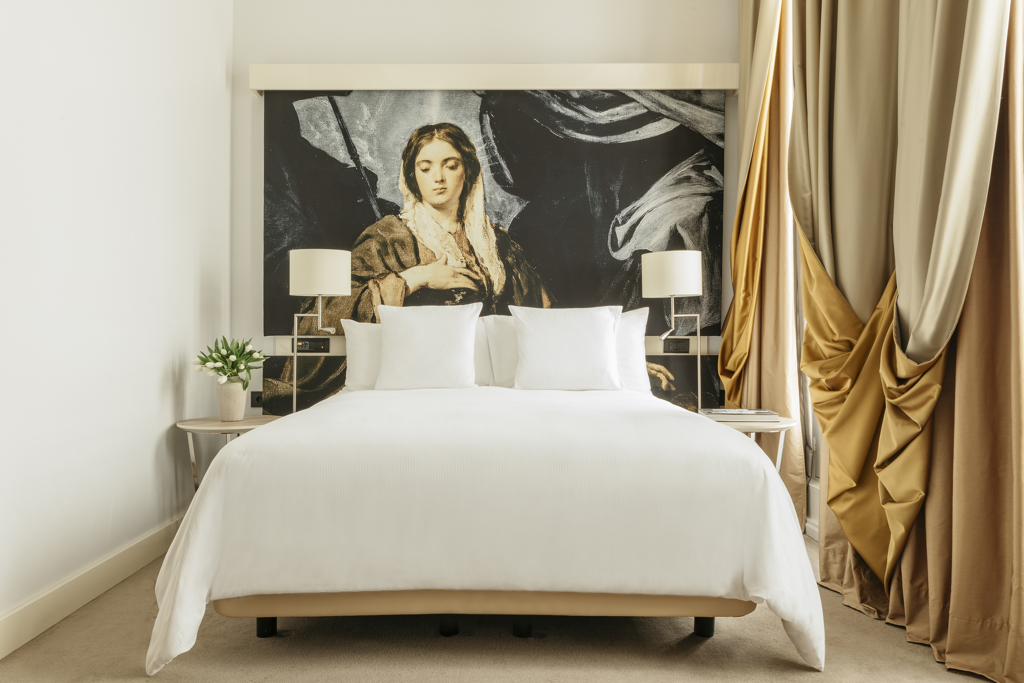 Elegant hotel room with white bed, classical art mural, and gold curtains.