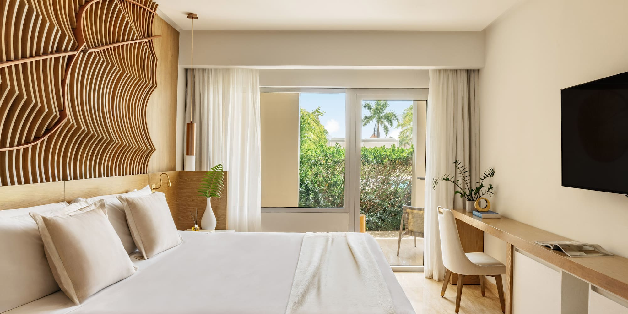 Modern hotel bedroom with wooden headboard, desk, and balcony with tropical view.