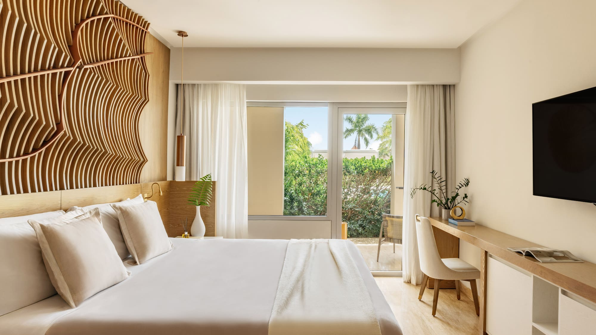 Modern hotel bedroom with wooden headboard, desk, and balcony with tropical view.