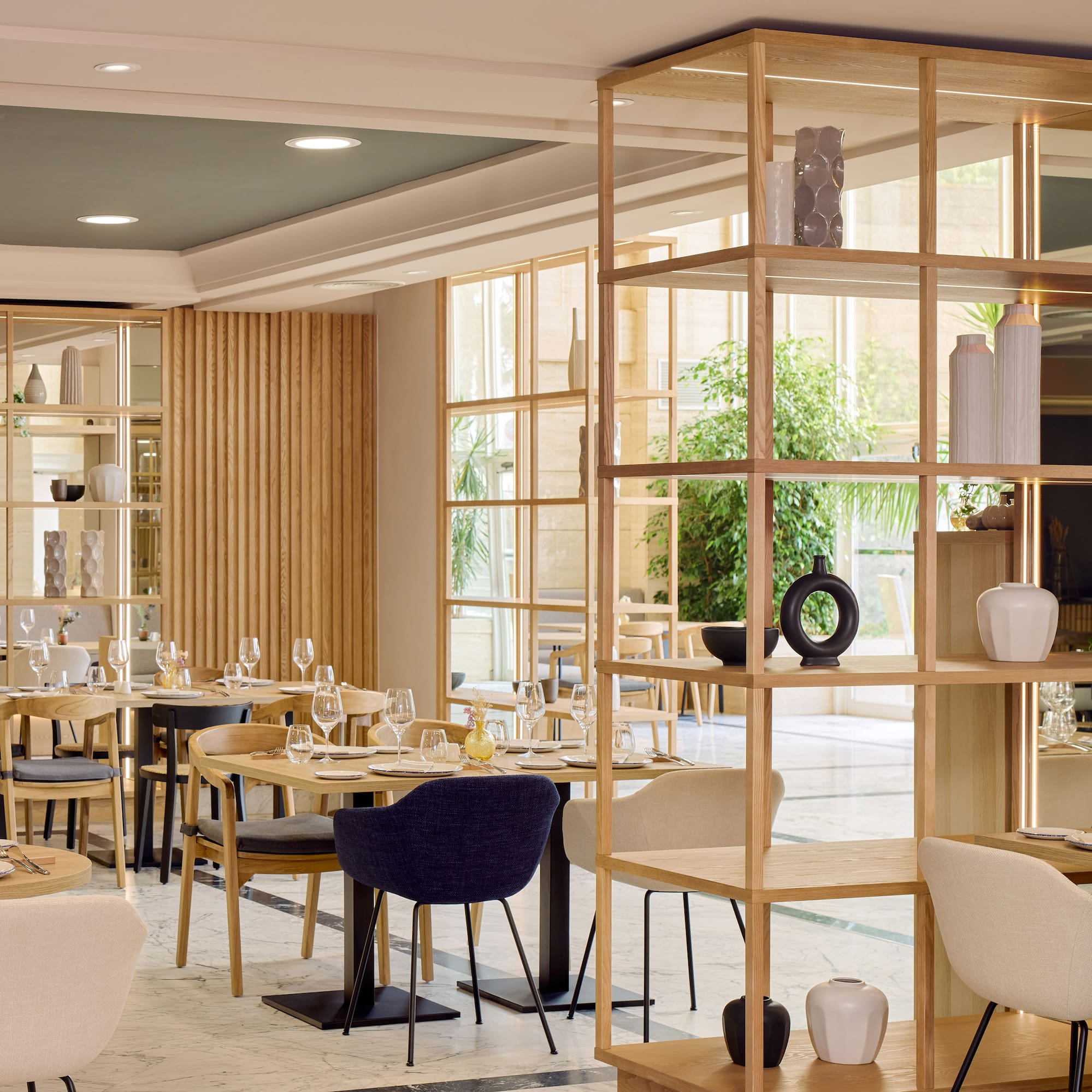 Restaurante moderno, mesas puestas, estanterías de madera y ventanales.
