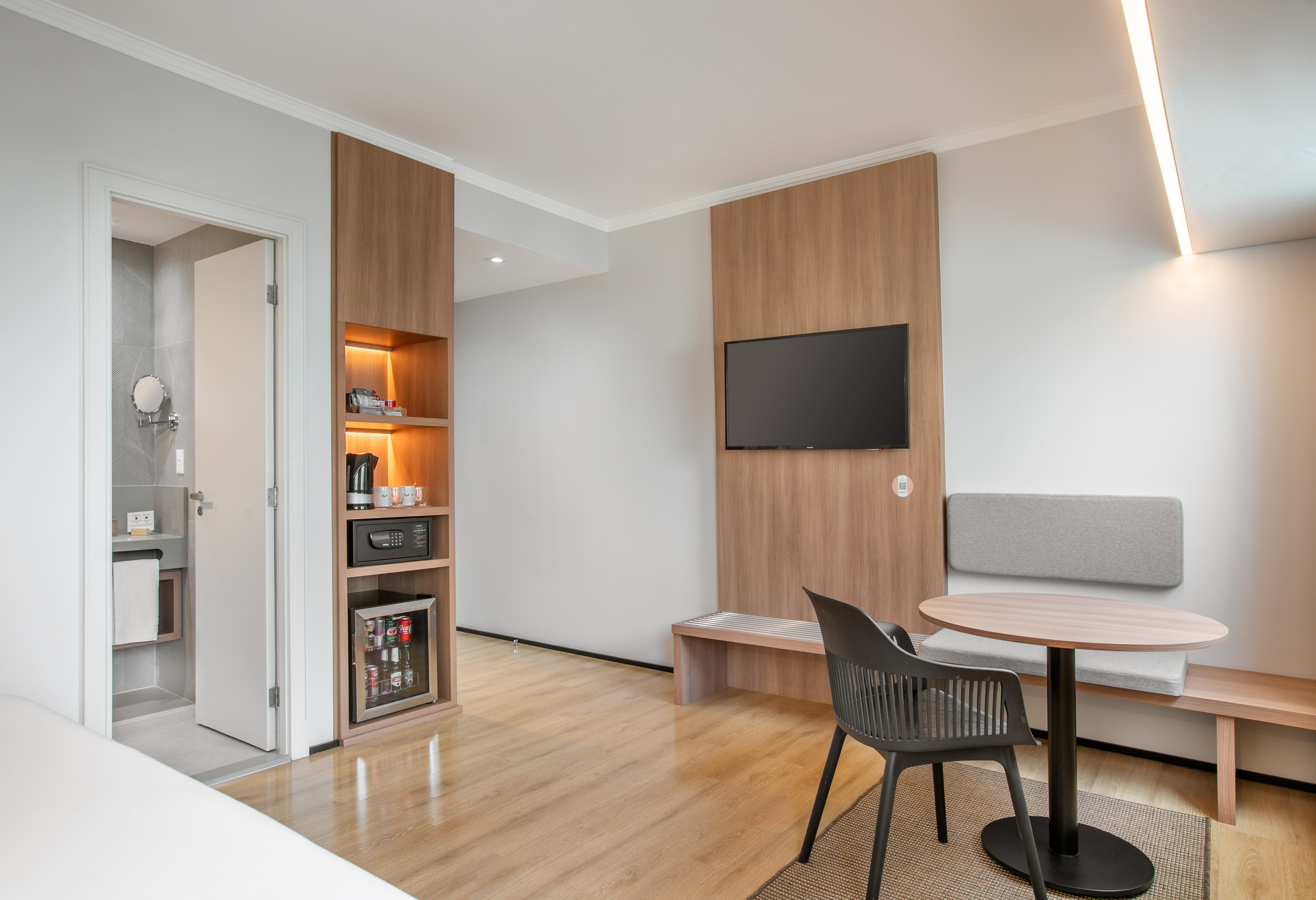 Modern hotel room with TV, wood paneling, minibar, coffee station, and seating.