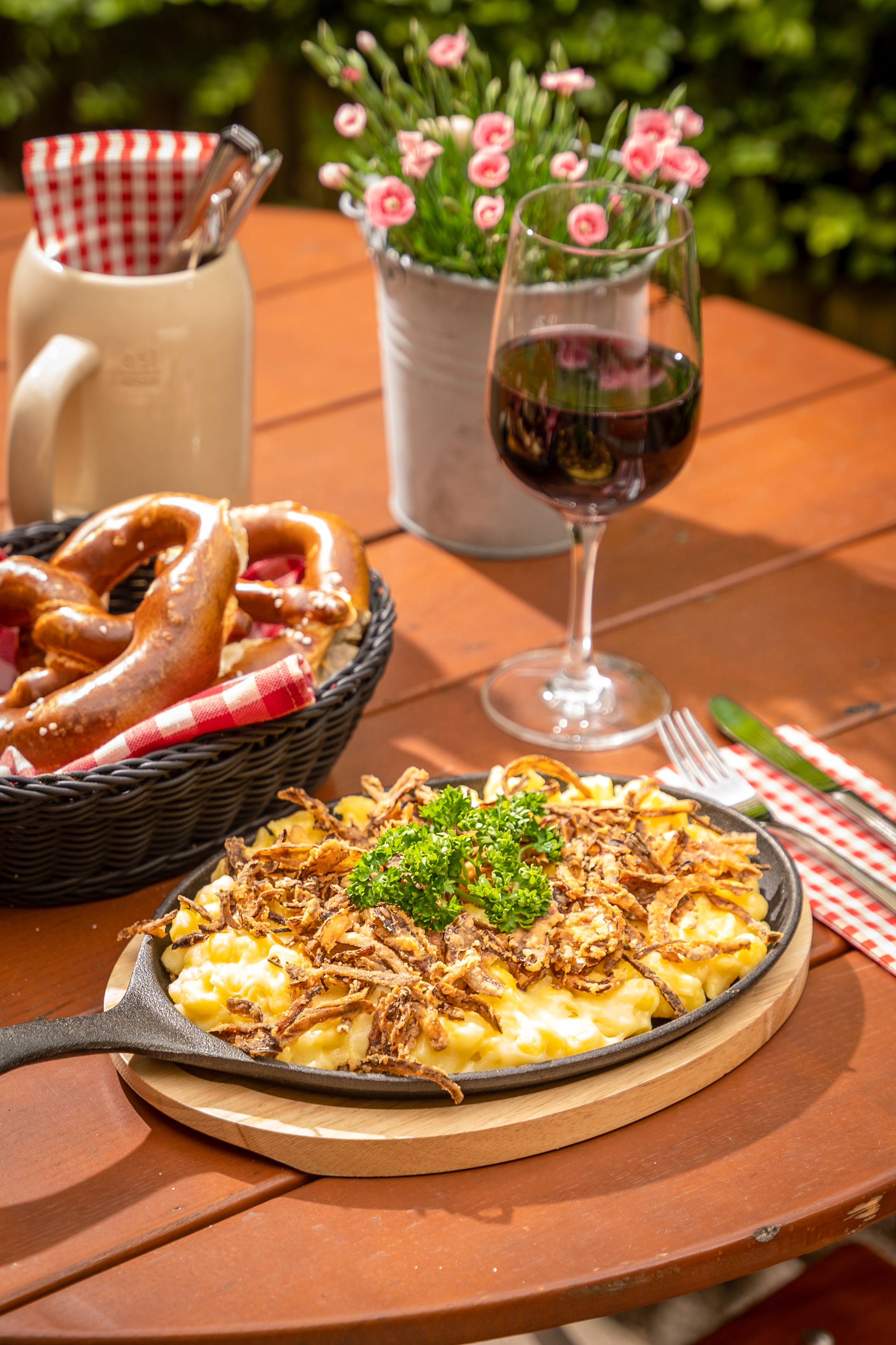 Bavarian Käsespätzle with fried onions, pretzels, and red wine on an outdoor table.