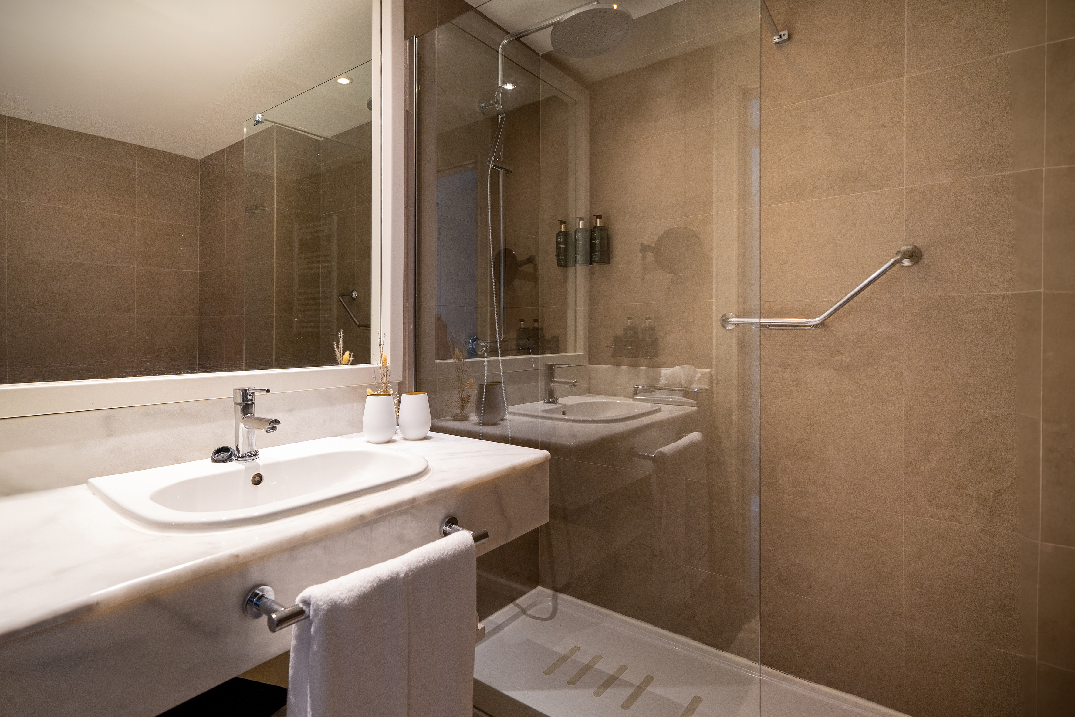 Modern hotel bathroom with marble vanity, sink, walk-in shower, and grab bar.