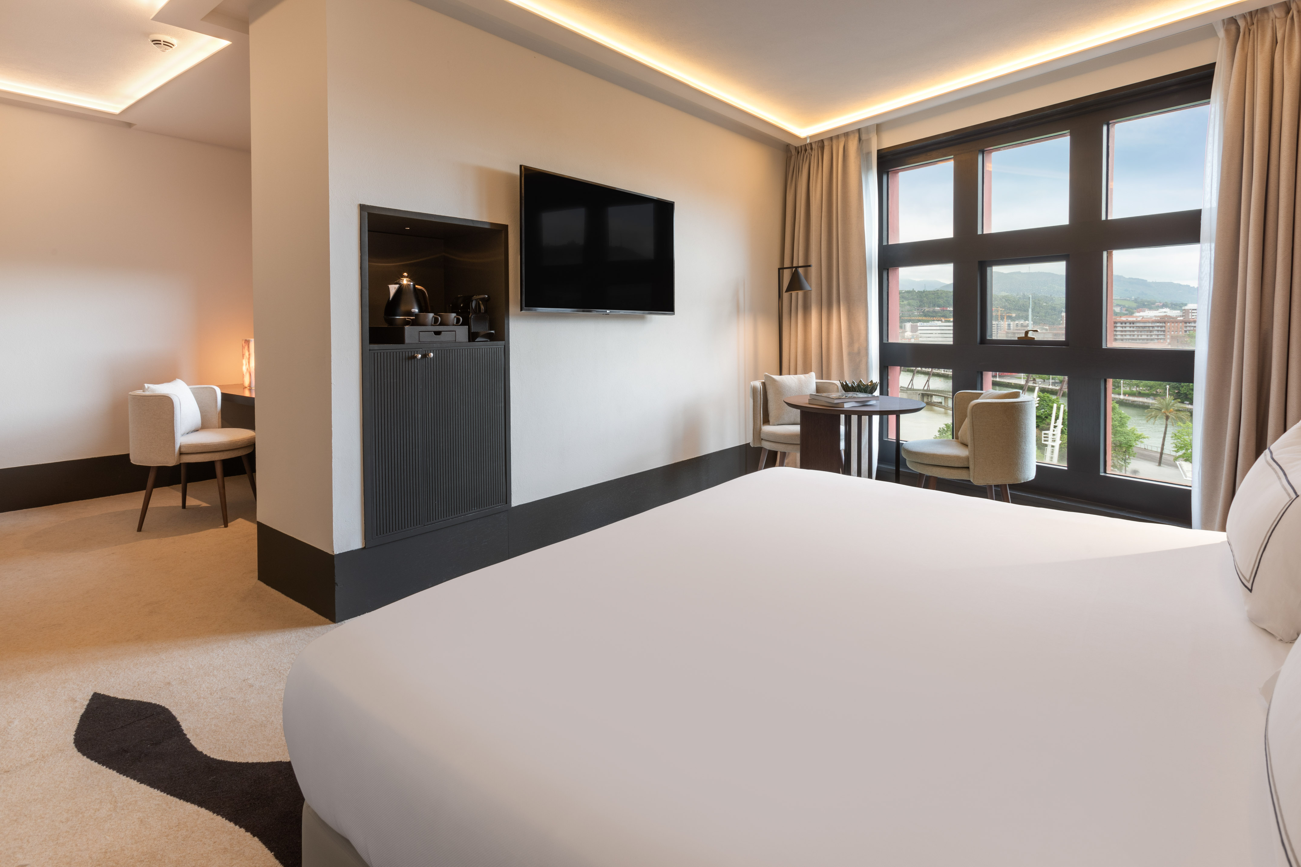 Modern hotel room with king bed, wall TV, minibar, and panoramic city view.