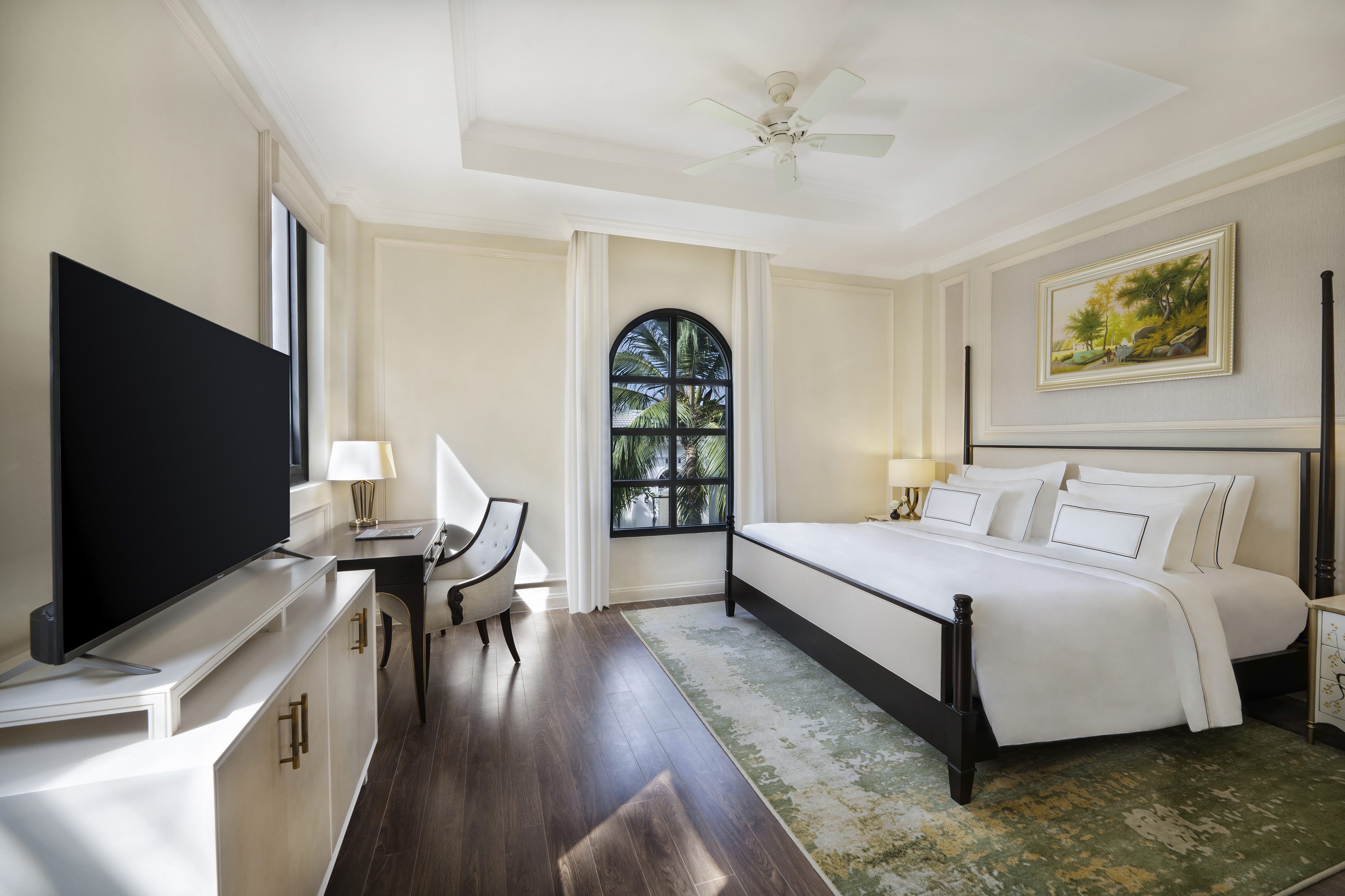 Modern hotel bedroom with king bed, TV, desk, arched window, palm trees.