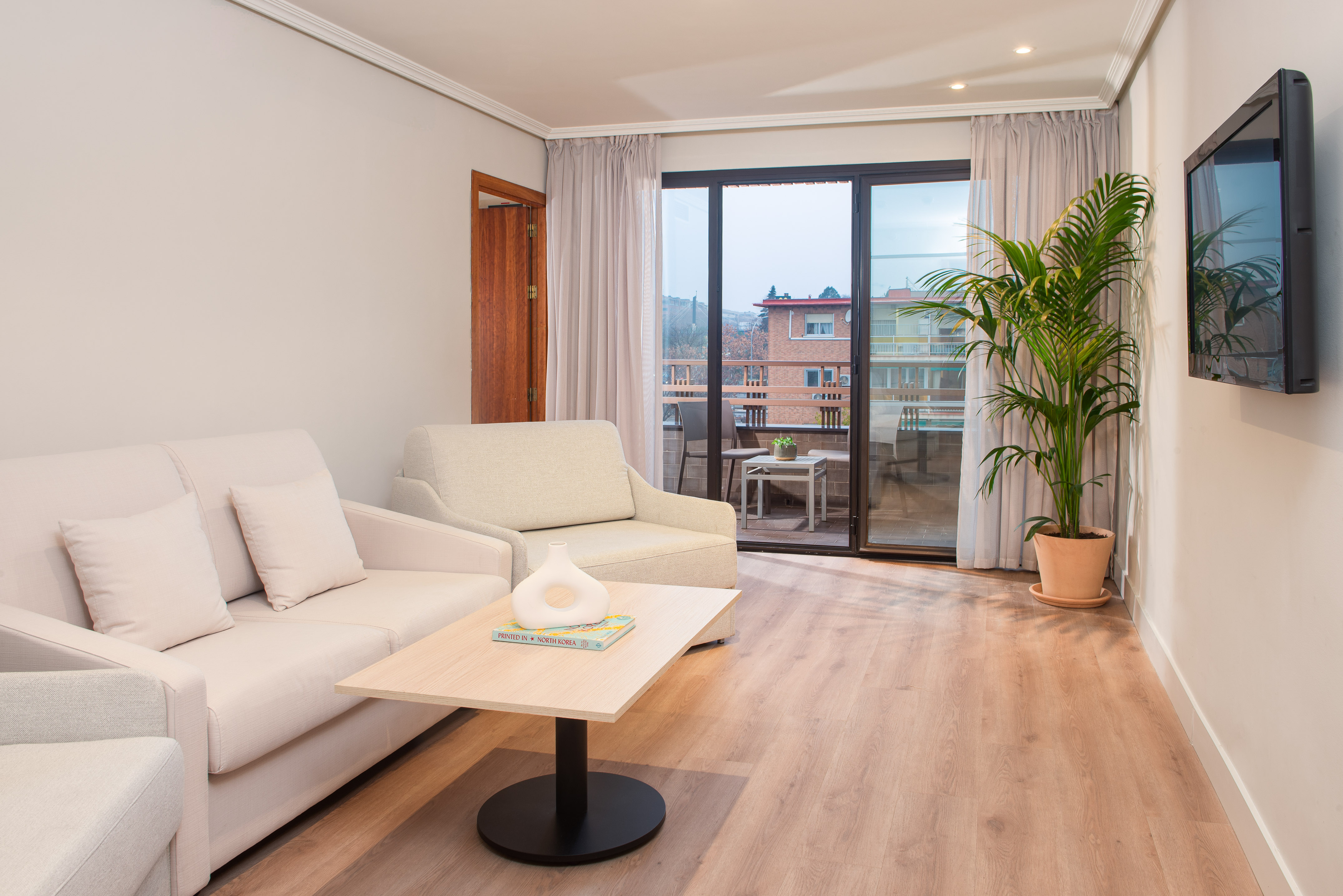 Modern hotel suite living room with sofa, coffee table, balcony, plant, and TV.